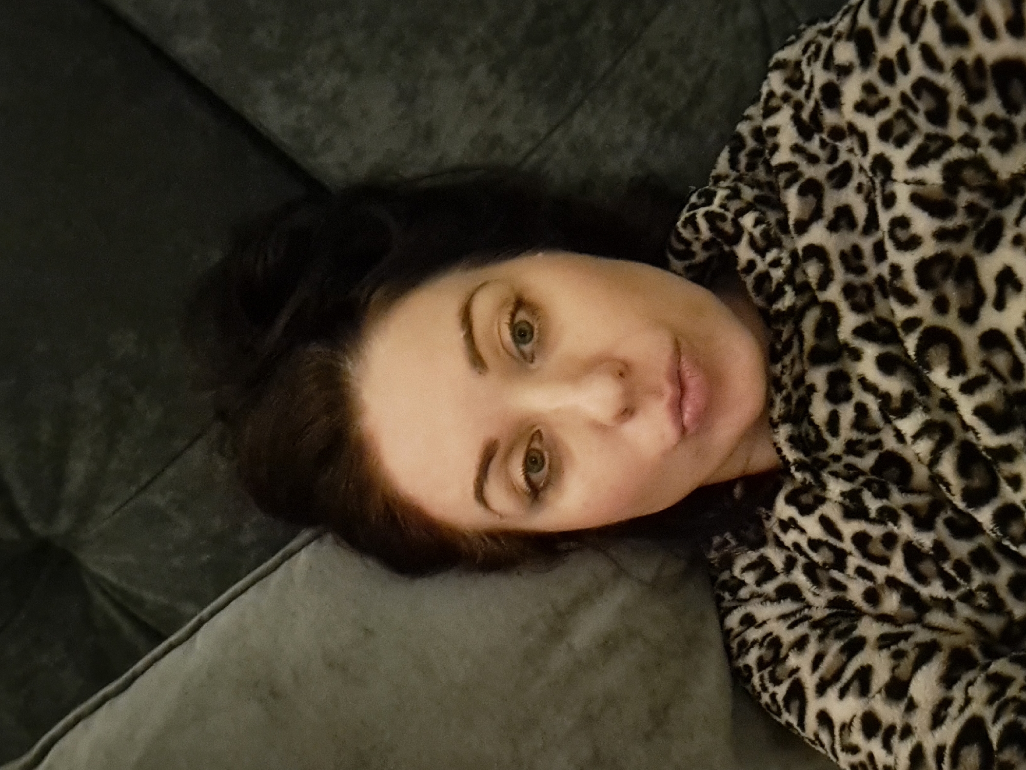 The image shows a close-up, selfie-style shot of a woman lying down, possibly on a couch, with her head angled towards the camera. She has dark hair pulled up and is wearing a fuzzy leopard-print garment. Her eyes are open, and her lips are pursed. The background appears to be a dark, textured surface, possibly upholstery. There is no visible indication of the city of Irkutsk, Russia, or any specific urban environment.