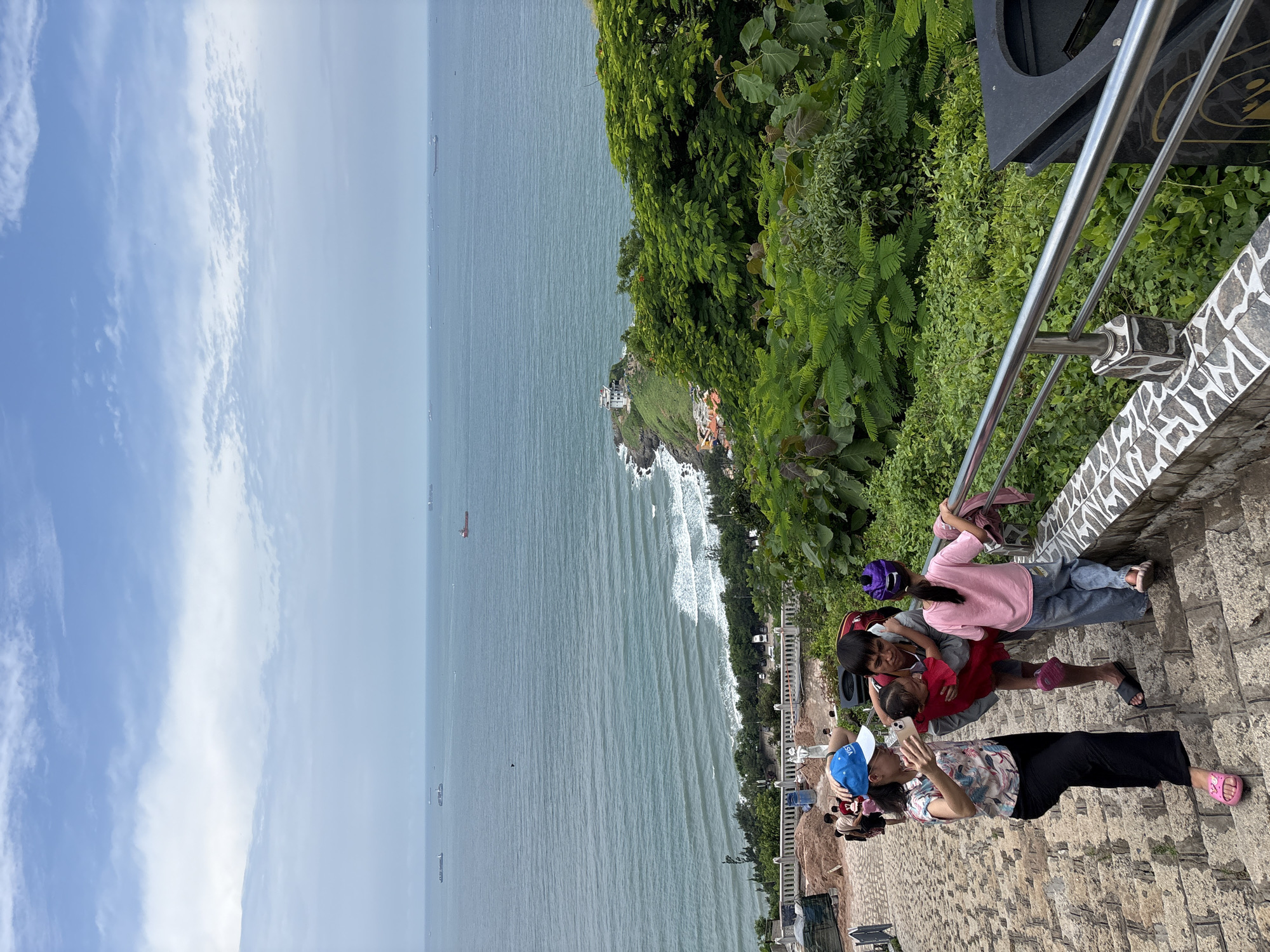 This is a vertical, eye-level shot of a stone staircase on a hillside overlooking the ocean. A family is ascending the stairs. A woman in a patterned blouse and black pants, wearing a blue baseball cap and pink sandals, takes a photo with her phone of a man holding a baby in a red dress. A young girl in a pink t-shirt and jeans walks ahead of them, holding onto a railing. The railing is made of metal and stone. Behind the people, lush green foliage spills over the edge of the staircase. The ocean is visible in the background, with several cargo ships dotting the horizon under a partly cloudy sky. In the distance, a white lighthouse sits on a rocky outcropping. Construction materials are visible on the left side of the staircase, suggesting ongoing work.