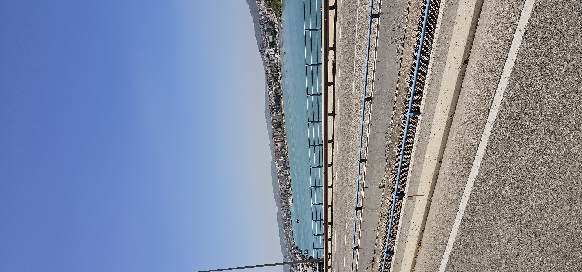 The image captures a vibrant coastal scene from the perspective of a bridge, overlooking a bay or strait in Algeciras, Spain. The primary orientation, once corrected from the provided vertical crop, reveals a panoramic view under a clear blue sky.
In the immediate foreground and extending into the midground on the right, there is a section of asphalt road or bridge surface. It features a distinct white painted line running parallel to concrete barriers and a blue metal guardrail or safety barrier, which separates the road from the water below. Another, shorter section of blue guardrail and concrete structure is visible further right, suggesting a wider bridge or multiple lanes. A thin, dark cable or wire extends upwards from the bottom of the frame, hinting at the bridge's construction.
Beyond the bridge structure, a vast expanse of calm, turquoise-blue water dominates the midground. The water appears still, reflecting the brightness of the sky. In the distance, dotting the water, a few small, dark shapes suggest boats or buoys.
Across the water, the background reveals a dense urban landscape stretching along a hilly coastline. Numerous buildings, varying in height and style, form a continuous strip of development, likely a city or town, nestled against the natural slopes of the land. The architecture appears contemporary, indicative of a bustling coastal community. Further back, the hills recede into a hazy, lighter blue, blending with the sky.
The left side and upper portion of the corrected image are dominated by a vast, cloudless, bright blue sky, indicating excellent weather. The lighting suggests it is daytime, likely late morning or early afternoon, with ample sunlight illuminating the entire scene. There are no visible people or specific activities taking place, and no discernible text within the frame. The overall impression is one of a serene yet active coastal urban environment.
