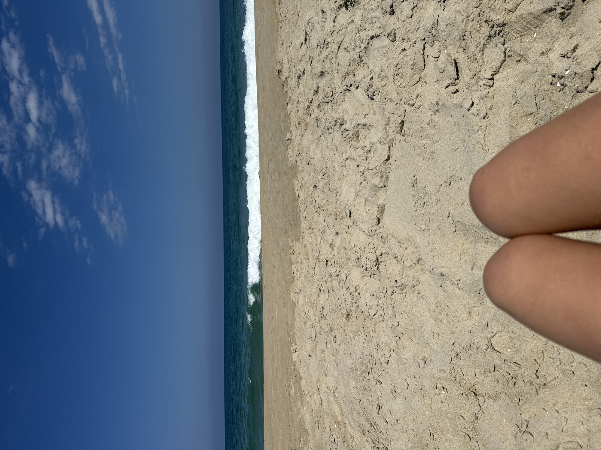 A low-angle, eye-level shot of a beach in Nags Head, United States of America, features a clear blue sky with scattered white clouds above a tranquil ocean with a gentle wave breaking. The foreground consists of sandy beach with textures of the sand visible, and the lower part of the frame shows a person's bare legs.