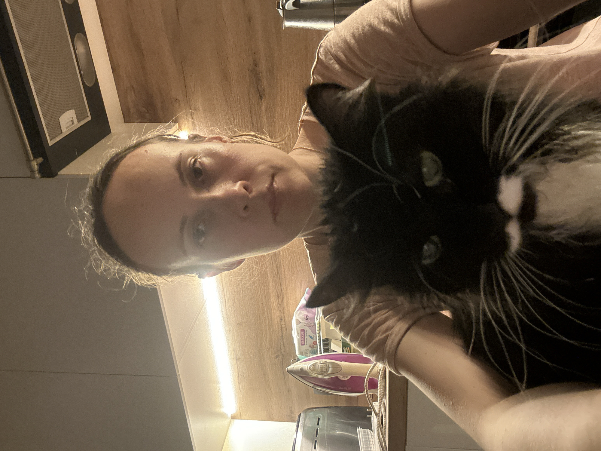 A mid-shot, eye-level, indoor, selfie-style photograph shows a person holding a black and white cat in their arms. The person, a woman with her hair pulled back, is wearing a light pink t-shirt. She is looking directly at the camera with a neutral expression. The cat is positioned in front of her, obscuring much of her torso. The cat has bright green eyes and prominent white whiskers. Behind them, a kitchen counter is visible with various items including an iron, a toaster, and a package of baby wipes. The background consists of wooden paneling and cabinetry, with a range hood mounted above. The lighting appears to be artificial and warm, casting shadows and highlights on the subjects and the environment. The overall scene suggests a casual, domestic setting.