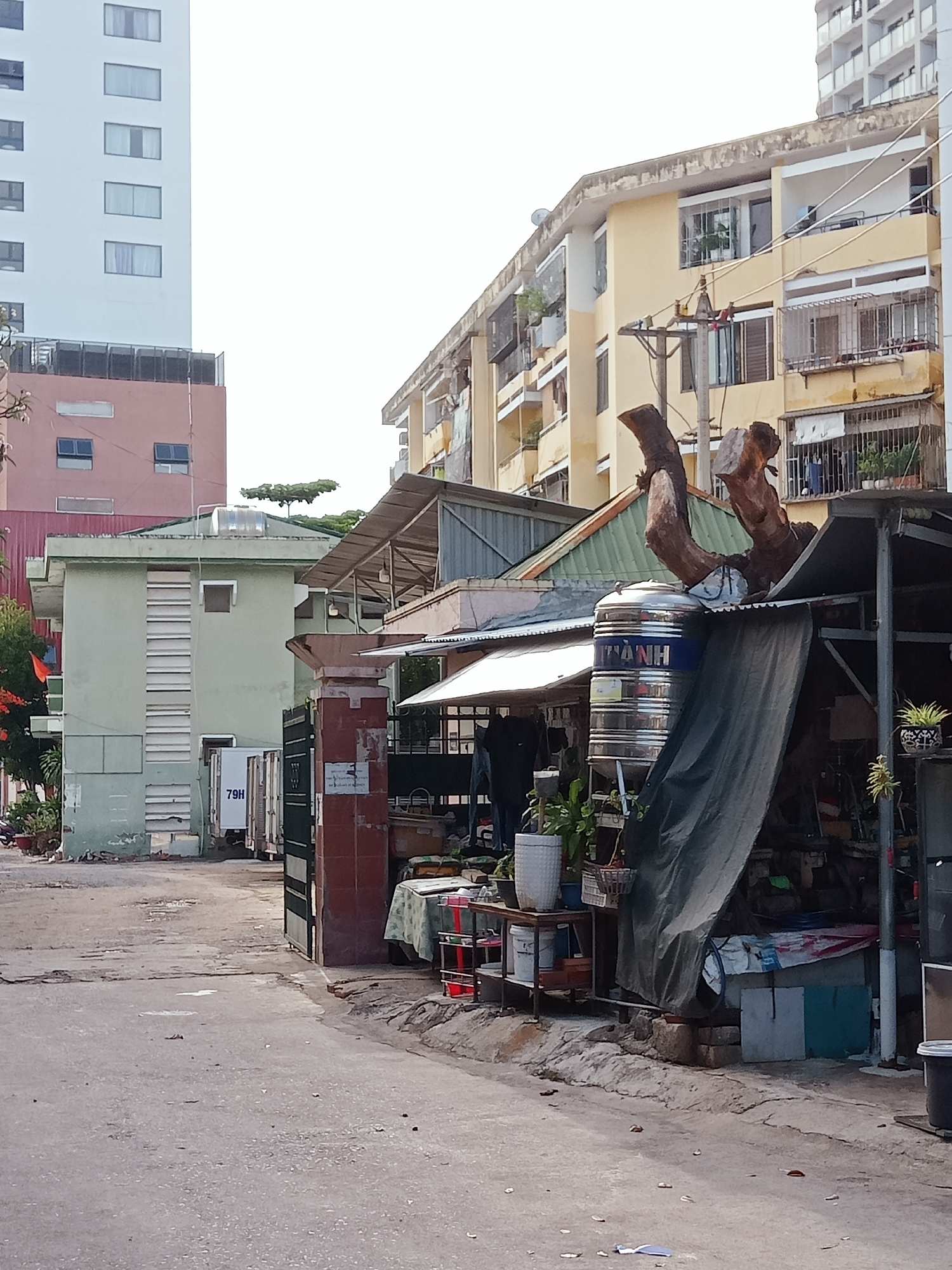 The image depicts a street scene in Nha Trang Ward, Vietnam. Buildings of varying heights and architectural styles line the street. A tall white building is visible in the background, partially obscured by a pink building. A lower, yellow building with balconies runs along the right side of the frame. In the foreground, a makeshift structure covered with a tarp sits on the right side of the street, with a water tank on top. Various objects, including plants and household items, are visible within and around the structure. A narrow, weathered street extends into the distance, with a small green building and a fenced area on the left. A sign reading "79H" is affixed to the green building. The street surface is cracked and uneven, with scattered debris.