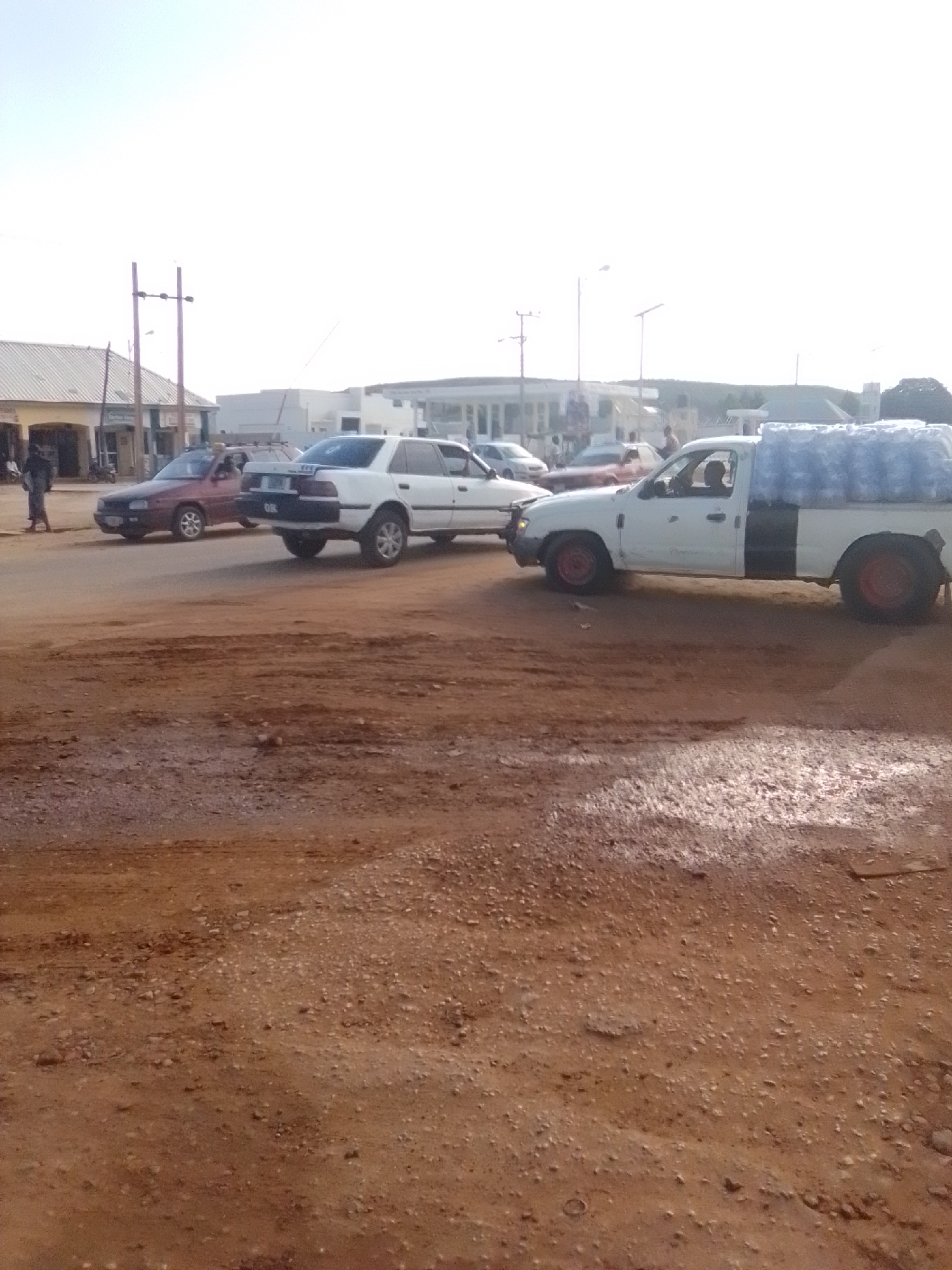 In Galadima, Nigeria, a white pickup truck filled with blue water jugs is parked on a dirt road. The truck has a black section on its rear passenger side and red rims on its back wheels. To the left of the truck, a white sedan is parked, followed by a red sedan. In the distance, several buildings line the street, including what appears to be a storefront. A person is walking on the left side of the road, and several other people are visible near the buildings. The sky is clear and bright, indicating daytime.
