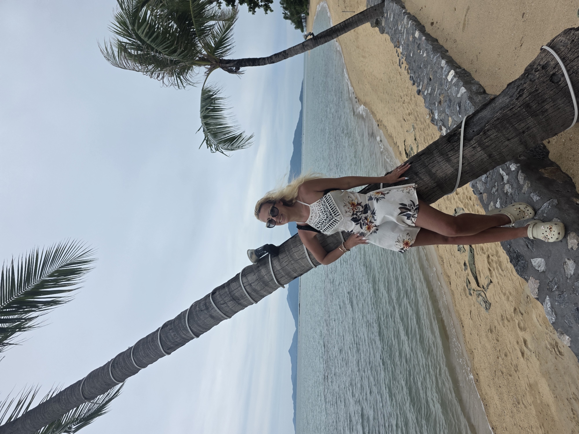 A blonde woman wearing a white floral romper and beige clogs stands leaning against a tilted palm tree on the sandy beach of Baan Bo Phut, Thailand. The ocean stretches out before her with distant islands on the horizon under a cloudy sky. A stone wall separates the beach from a path behind the woman.