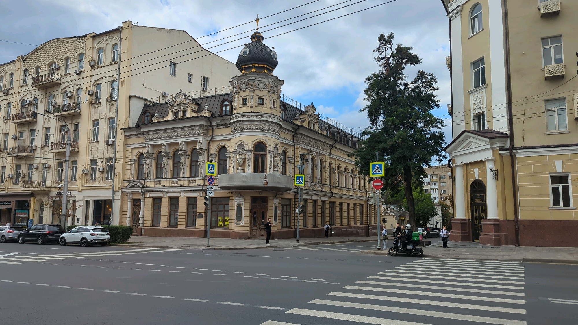 This image shows a street view in Rostov-on-Don, Russia, focusing on a distinctive historical building with ornate architecture. The building is painted in a pale yellow color and features a dark, bulbous dome topped with a spire and golden accents. It has multiple floors with arched windows and decorative detailing, including balconies with wrought-iron railings on the left side of the image. In front of the building, there is a crosswalk on a paved road.
Several vehicles are visible on the street: a white SUV, a dark sedan, and a white car parked in a line. A moped with a green delivery box is also present, with two people on it, one appearing to be the driver and the other a passenger wearing a helmet. Three individuals are standing on the sidewalk near the moped, and another person is standing on the sidewalk in front of the historical building.
Traffic signs, including pedestrian crossing signs (blue squares with a white pedestrian symbol) and a red circular prohibition sign, are positioned at the intersection. The sky is partly cloudy, with a mix of gray clouds and blue sky visible. A large green tree stands to the right of the historical building. The overall scene depicts an urban environment with a blend of historical and modern elements, likely a public street in the city center.