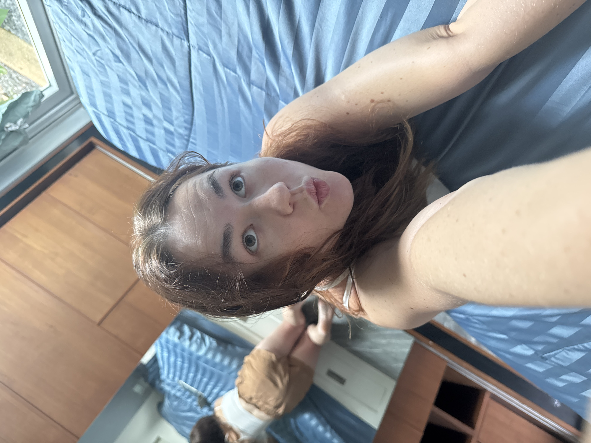 The image shows a woman lying on a blue, striped bed. She is wearing a white tank top and brown shorts. Her reflection is visible in a mirror to her left, where she is also seen wearing the same outfit and is in a similar pose. The room appears to be a bedroom with wooden furniture, including a closet. The overall impression is that of a casual, personal moment captured in a bedroom setting. The provided context states the location is Rawai, Thailand, suggesting this bedroom is located in that area.