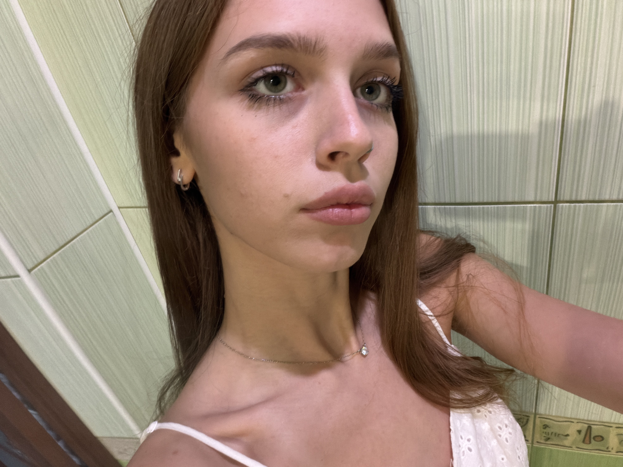 This media file is a close-up, waist-up portrait of a young woman with long, brown hair. She is facing slightly to her right, with her gaze directed upwards and to the right, away from the camera. She has fair skin, light green eyes, and her eyebrows are dark and well-defined. Her lips are parted slightly, and she appears to be wearing light makeup, with noticeable mascara on her eyelashes.
She is wearing a delicate silver chain necklace with a small, clear pendant. Her clothing is a white, sleeveless top with intricate embroidered details along the neckline and strap. A subtle shadow is cast on her face from an unseen light source.
The background consists of pale green, tiled walls with vertical lines. To the left of the frame, there are some vertical wooden or paneled elements in darker shades, suggesting an indoor setting, possibly a bathroom or a room with tiled walls. The lighting appears to be artificial and relatively dim, creating a soft ambiance. No specific time of day or weather conditions can be determined. There is no visible text in the image.