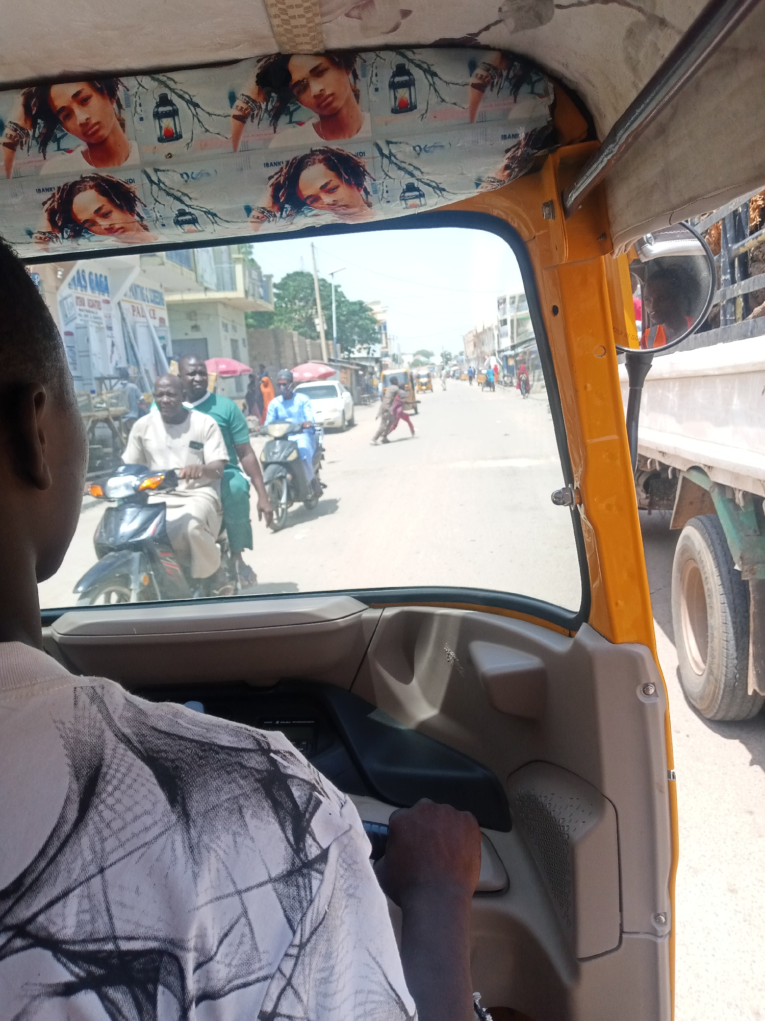 An interior view, from the perspective of a passenger, captures the scene of a bustling city street. The foreground is dominated by the interior of a yellow motorized rickshaw, with the driver's shoulder and the top of his head visible on the left side of the frame. The roof lining of the rickshaw is decorated with repeating images of a young man's face and small lanterns. A side mirror reflects the driver wearing an orange vest.
Outside the rickshaw, the street is filled with activity. Two men on a motorcycle are seen in the middle ground, with the driver wearing a light-colored tunic and the passenger in a green shirt. Another motorcycle, ridden by a person in a blue shirt, is to their right. In the background, a white truck is visible on the right, partially obscuring a busy street lined with buildings and people. Several other vehicles, including a yellow auto-rickshaw, are also present on the street. The overall impression is one of a lively urban environment.