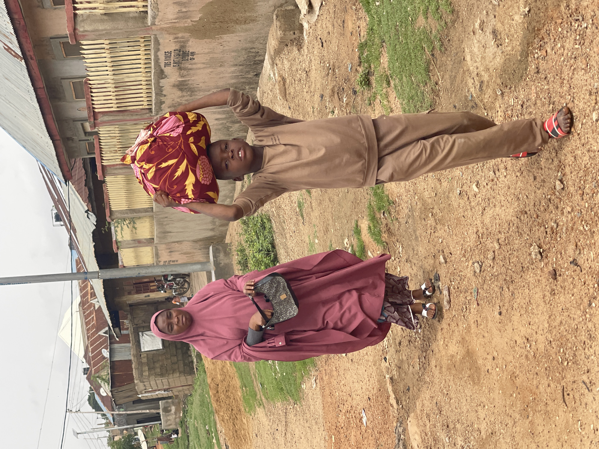 Two people stand on a dirt path in Unguwar Gabas, Nigeria. A young person in brown pants and a long-sleeved brown shirt carries a colorful bundle on their head. A woman in a reddish-purple hijab holds a small handbag. They are standing in front of a building with a corrugated metal roof and a white sign with text on it. Behind them are other buildings with corrugated metal roofs. Grass grows on the side of the dirt path.