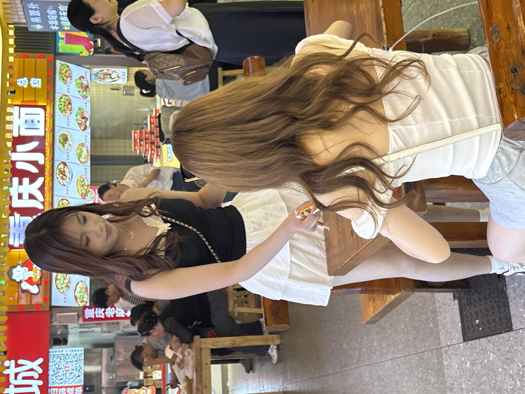This image captures a vibrant indoor or semi-outdoor food court or casual restaurant setting, likely in Yuzhong District, China. The scene is brightly lit, suggesting daytime or evening with strong artificial lighting.
In the foreground, two women are seated at a wooden table. The woman on the left has long, dark brown hair and wears a black top with lace details and a white pleated skirt. She holds what appear to be chopsticks and has a relaxed expression. The woman on the right, shown from behind, has long, wavy light brown hair and wears a white strapless or corset-style top with grey shorts. A white strap, possibly from a shoulder bag or her clothing, is visible over her back.
Several other people are visible in the background, both seated and standing, suggesting a busy environment. One woman in the upper background wears a white shirt and carries a brown patterned shoulder bag. The setting features rustic wooden tables and chairs.
The background is dominated by large, illuminated menu displays and neon signs. These displays showcase various food items, likely dishes served at the establishment.
Visible text on the signs includes:
- "城庆小面" (Chéng Qìng Xiǎo Miàn), which translates to "City Celebration Noodles" or "Chongqing Xiaomian," a popular local dish.
- "重庆老庄" (Chóngqìng Lǎo Zhuāng), meaning "Chongqing Old Village/Farmstead."
- "总店" (Zǒng Diàn), indicating it's the "Main Store" or "Headquarters."
- "牛肉" (Niú Ròu), meaning "Beef."
- Other phrases like "鼎源外" (Dǐng Yuán Wài), "水需现榨" (Shuǐ Xū Xiàn Zhà - "Water needs freshly squeezed," possibly for juice), and "健庙石价" (Jiàn Miào Shí Jià).
- A QR code is also visible on one of the signs, commonly used in China for payments or information.
The overall impression is that of a bustling eatery, typical of a food street or market in an urban Chinese setting.