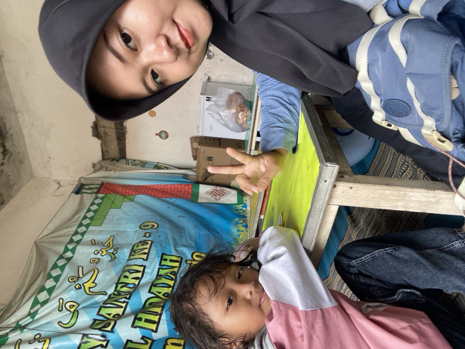 Two people are in the image. One is wearing a black hood, blue shirt, and light blue jacket. They are holding up three fingers. The other person is a child with brown hair, wearing a pink and white shirt. There is a table with a yellow top. The background includes a banner with text in a different language, a white refrigerator, and cardboard boxes. The room looks cluttered and aged.