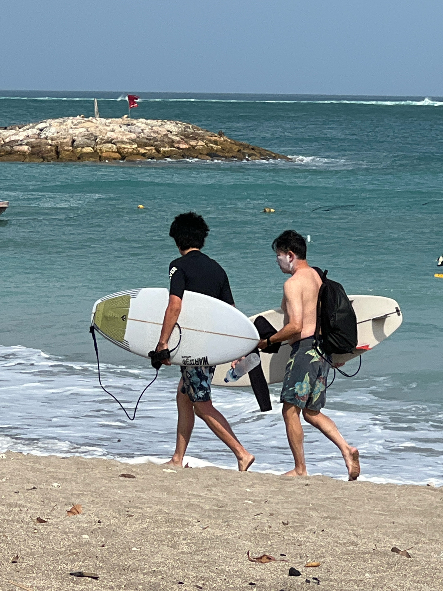 Two individuals, presumably surfers, walk along the shoreline of Kuta Beach, Indonesia, carrying surfboards. The person on the left, wearing a black wetsuit and patterned swim trunks, holds a white surfboard with green accents. The person on the right, shirtless and wearing patterned swim trunks, carries a white surfboard and a black backpack. They are walking from left to right. The ocean is calm with gentle waves lapping at the sandy shore. In the background, a rocky outcrop with a red flag is visible, along with the expanse of the ocean and a clear sky. The sand is a light brown color and scattered with a few dry leaves and debris. The overall scene depicts a casual beach activity.