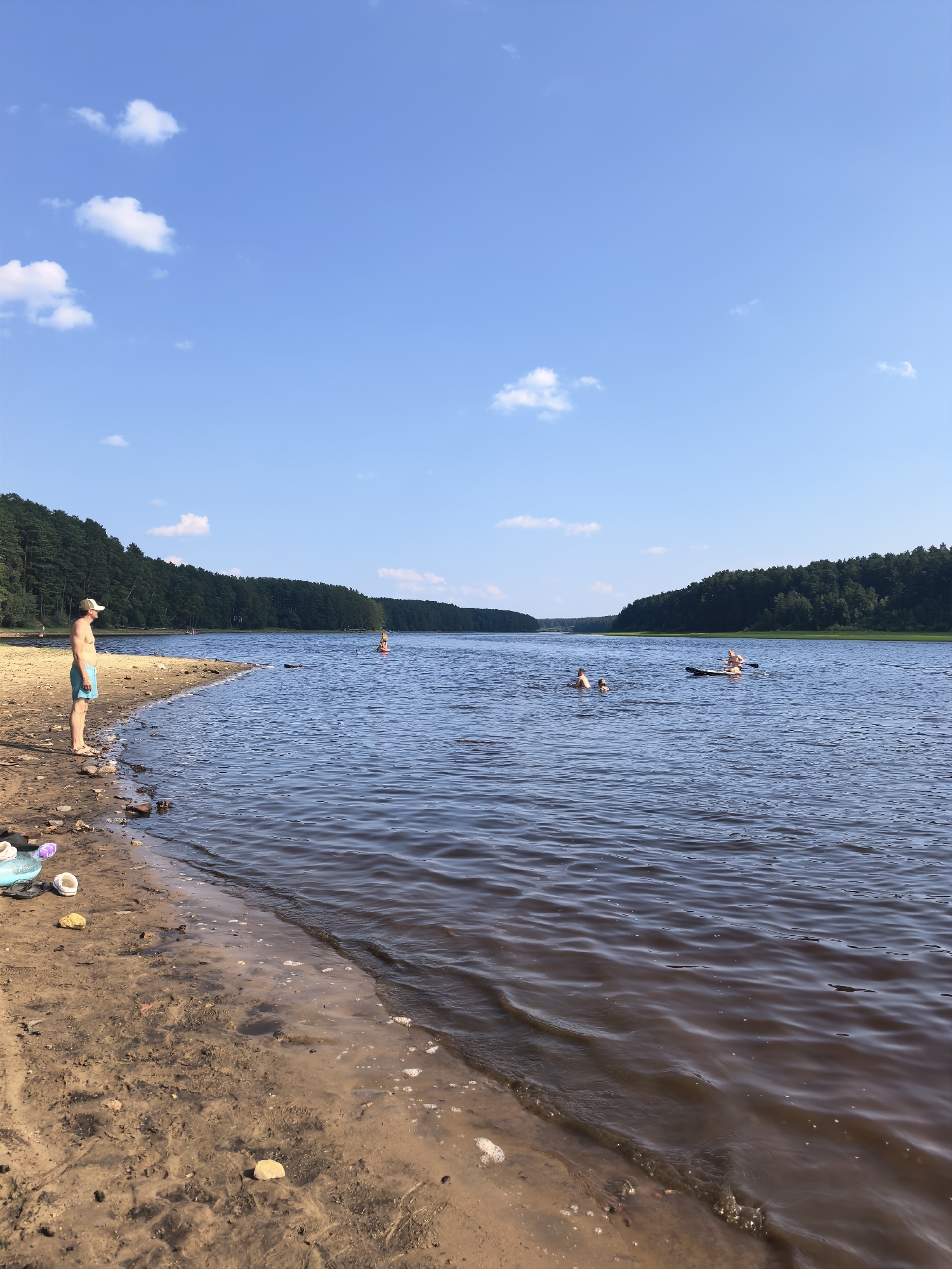 This media file depicts a serene summer day at a lake in Sysert, Russia. The scene is dominated by a wide expanse of calm, dark water, reflecting the bright blue sky dotted with fluffy white clouds. A sandy shore in the foreground is lined with scattered rocks and some discarded footwear, including flip-flops and sandals, suggesting recent human activity.
On the left side of the frame, a man wearing blue swim trunks and a white cap stands on the beach, looking out at the water. He appears relaxed, enjoying the view.
Further out in the lake, several people are engaged in recreational activities. Two individuals are swimming, while another person is on a stand-up paddleboard. A bright orange buoy is visible in the distance, marking a point in the water.
The background is a lush, dense forest that stretches along the far bank of the lake, creating a natural and tranquil setting. The overall impression is one of a peaceful summer day, with people enjoying the water and the natural beauty of the Russian landscape. The time of day appears to be late afternoon or early evening, given the soft lighting. There is no visible text in the media file.