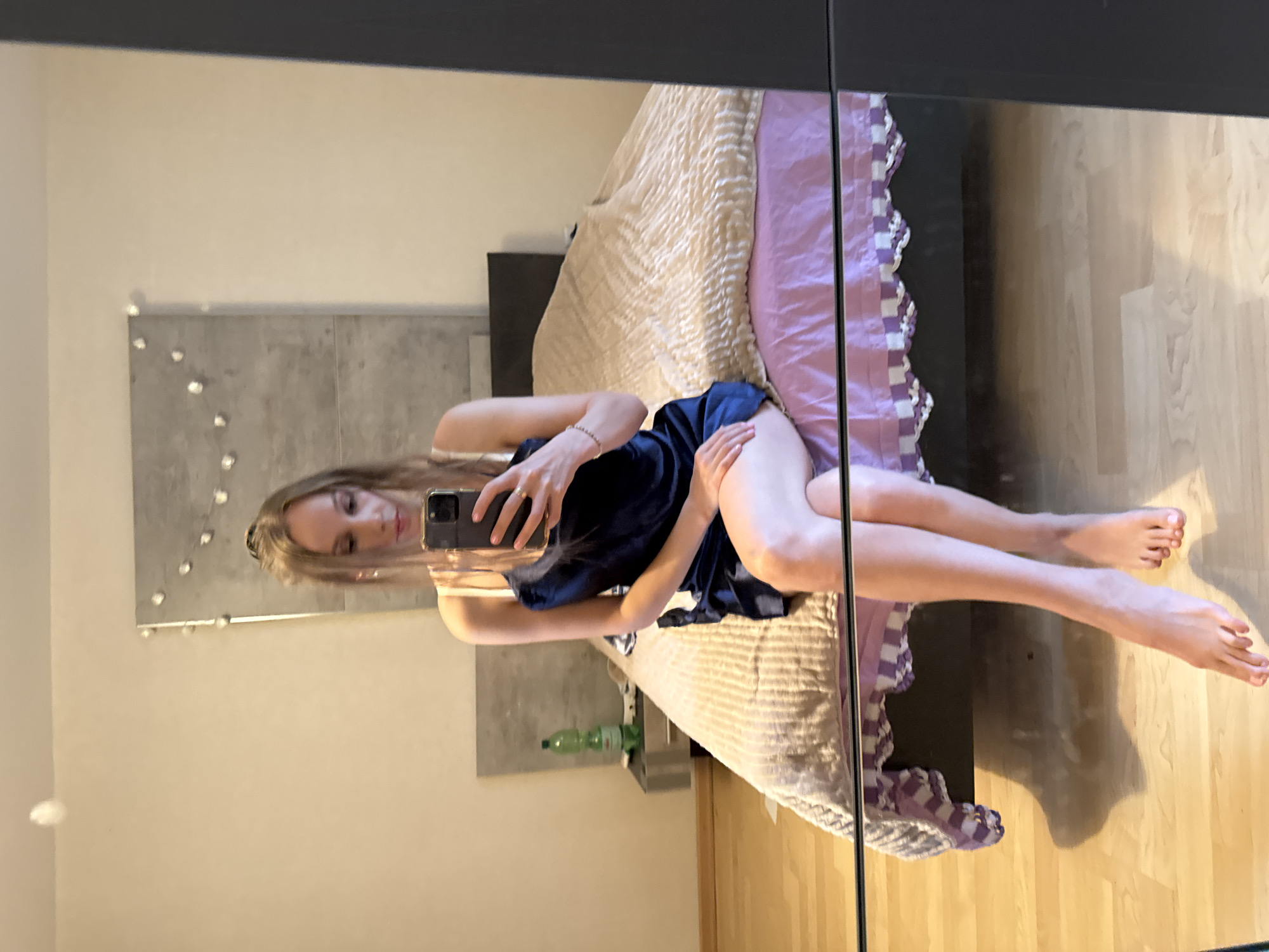 A young woman with blonde hair pulled back in a ponytail is sitting on a bed in a bedroom, taking a mirror selfie with her phone. She is wearing a dark blue slip dress and is looking down at her phone. The bed is covered with a textured beige blanket and a pink sheet. In the background, there is a bedside table with a bottle of water and a lamp. The room has light-colored walls and wooden floors. The reflection in the mirror shows the woman's full body and the surrounding room. The image is taken from a slightly high angle.