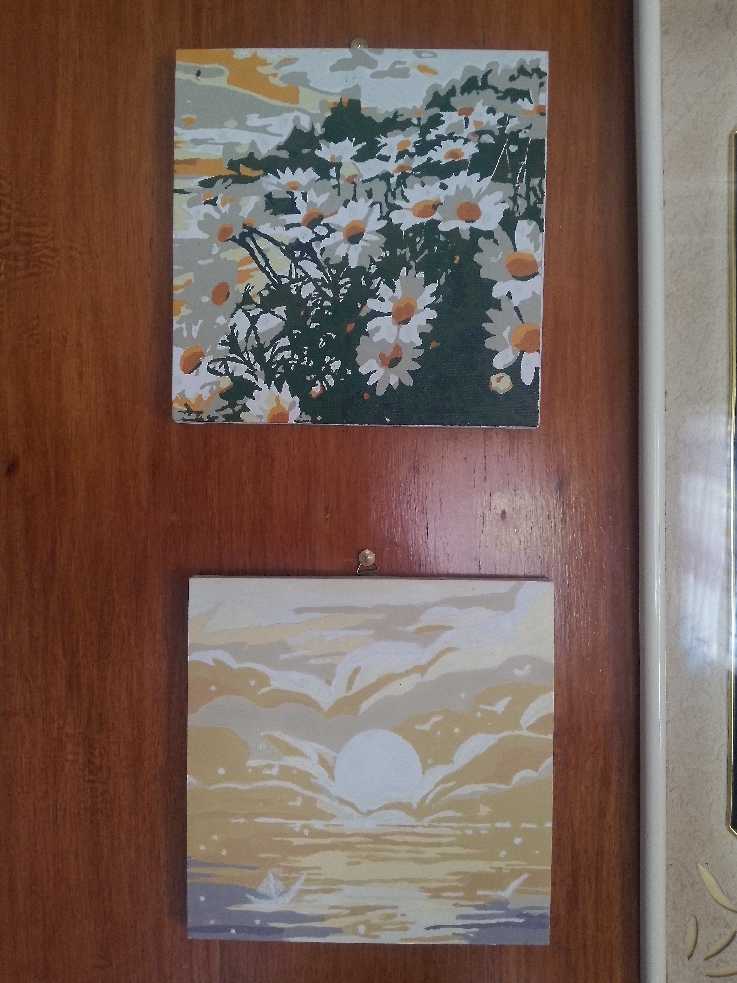 Two square paintings hang on a wooden panel. The top painting depicts a field of daisies under a cloudy sky. The bottom painting shows a sunset over the ocean with a paper boat. The wooden panel appears to be part of a door or cabinet. A white pipe is visible along the right edge of the image. This scene is not necessarily located in Davao City, Philippines.