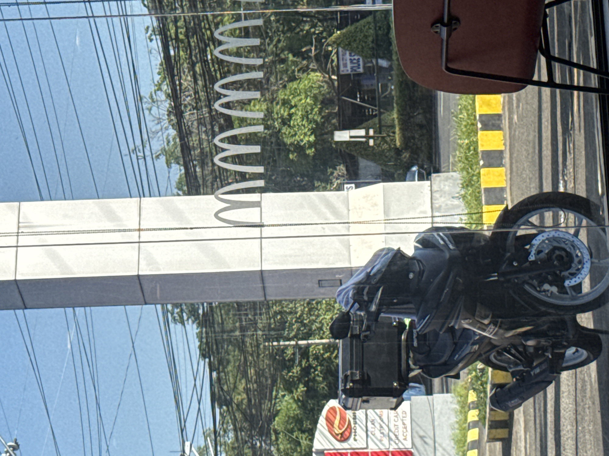 The image, when rotated 90 degrees clockwise, displays a bright, sunlit street scene viewed through a glass window, likely from inside a McDonald's restaurant in Davao City, Philippines. The foreground features a series of stylized "M" shapes, characteristic of the McDonald's logo, suggesting its location.
Beyond the window, a clear blue sky is visible, with numerous black utility wires crisscrossing horizontally. Abundant green foliage and trees line the background. Partially visible amidst the trees is a sign that reads "POLICE". On the right side, a black motorcycle, possibly a sport or commuter bike, is parked on a paved surface beside a curb distinctly marked with yellow and black stripes. The motorcycle's front wheel and disc brake are clearly discernible.
At the bottom center, a red logo featuring a flip-flop is visible, identifying "PHOENIX" (Phoenix Petroleum), indicating a nearby gas station or service area. Accompanying text on a sign partially reads "CLEAN RESTROOMS" and "CREDIT CARD ACCEPTED". A large, light-colored building or pillar runs vertically along the left edge of the frame, reflecting the sky. The scene captures a static moment of a typical urban landscape under bright, sunny conditions. No individuals are visible.