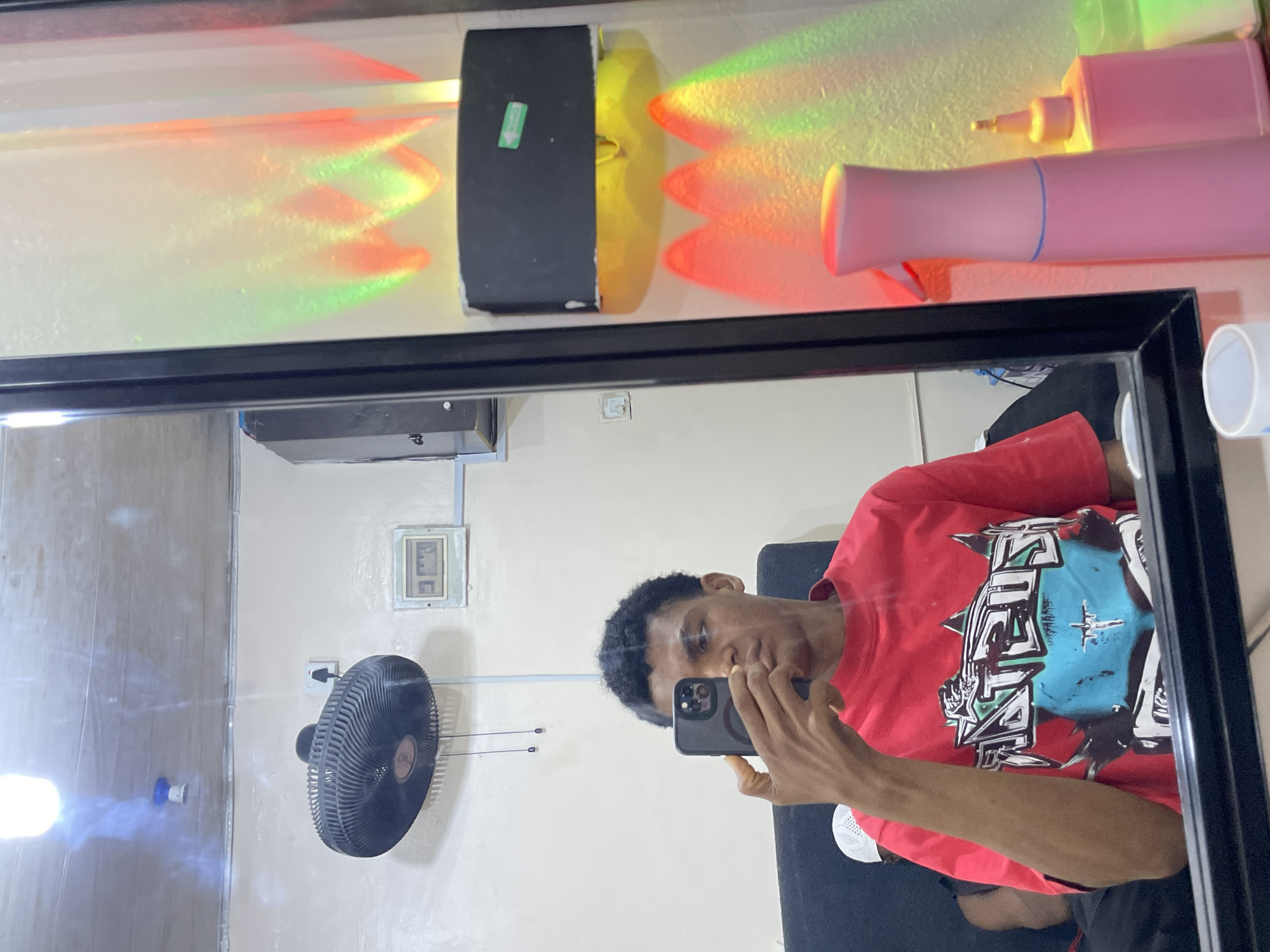 The image shows a reflection in a black-framed mirror. A young Black man wearing a red t-shirt with a colorful graphic is holding a smartphone, presumably taking the picture of himself. The background, reflected in the mirror, shows a room with light-colored walls. A black ceiling fan is mounted on the wall to the left. Other wall-mounted features include a small electrical outlet and a framed display of numerals or a gauge. The room appears to be lit by an overhead light source not visible in the reflection. On top of the mirror in the reflection, there is an odd, elongated light fixture with red and green colored rays. Also on the top of the mirror is a pink tube. It seems the picture was taken in a residence or private room in Ado, Nigeria.
