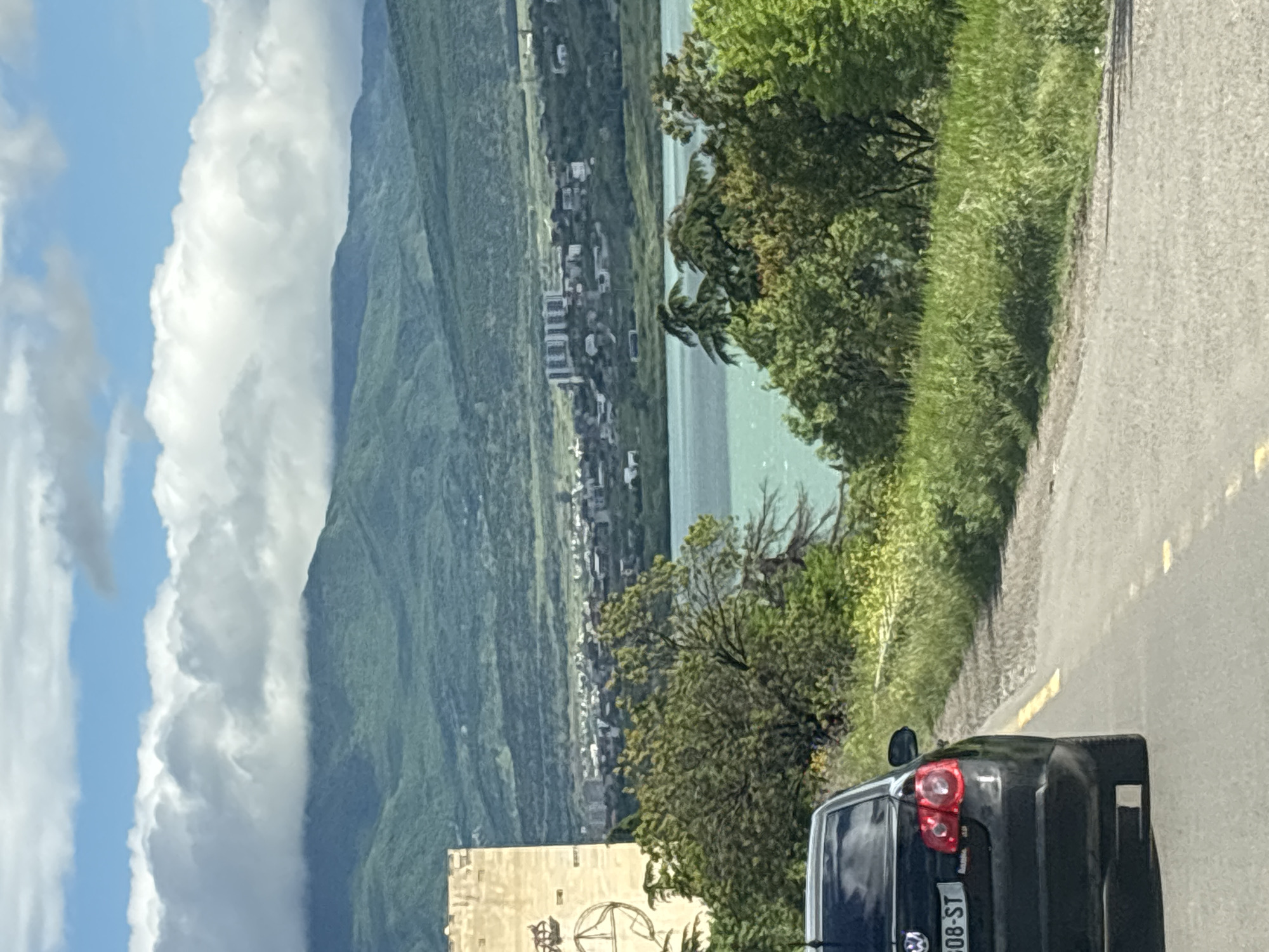 A medium shot depicts a road in Tbilisi, Georgia. To the left, mountains rise alongside a river, with buildings visible on the river's edge. The sky is a mix of blue and white clouds. A car occupies the lower right of the frame, its rear lights and license plate visible. Trees and grassy vegetation line the road. A stone structure partially appears in the lower-left corner.