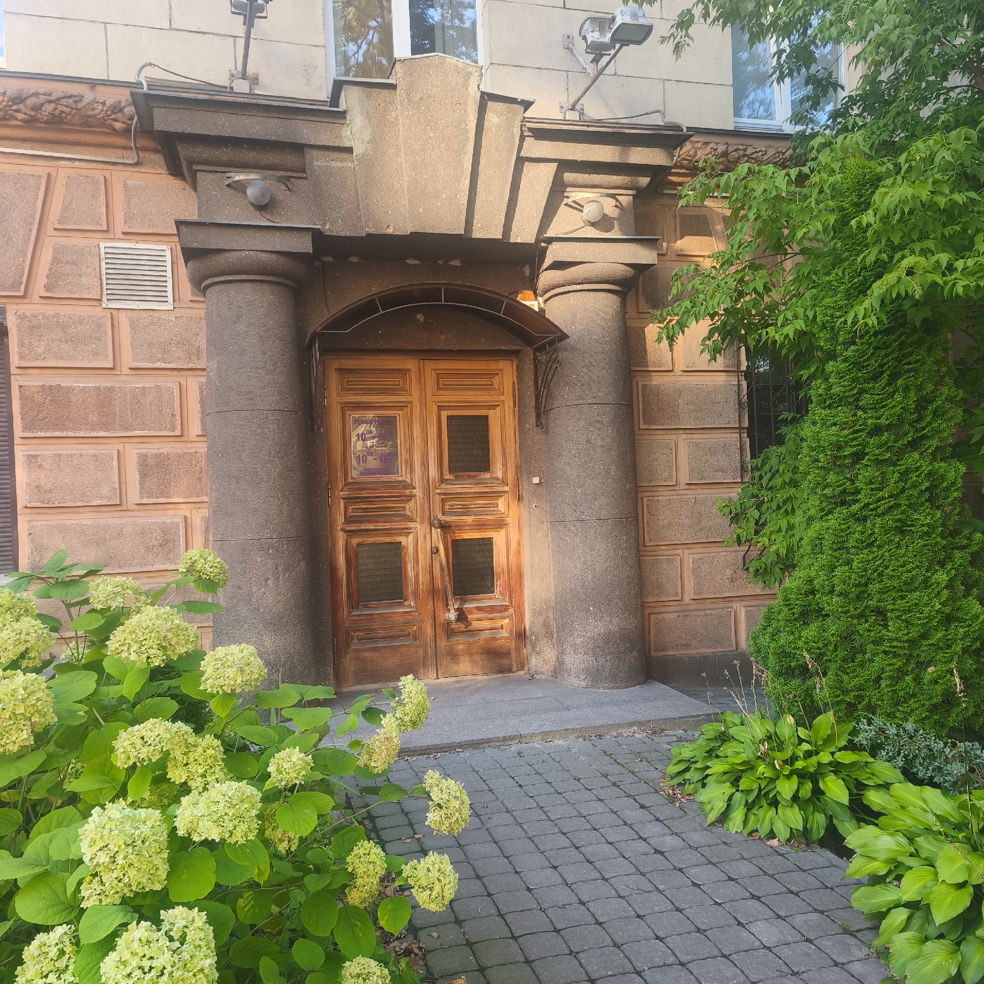 The image depicts the entrance to a building in Minsk, Belarus. The facade features a large, weathered wooden double door with glass panes. Flanking the door are two substantial stone pillars. Above the door, a small metal awning provides cover. To the left of the entrance, a white ventilation grate is visible on a wall with brick-like detailing. Lush green foliage, including flowering hydrangeas in the foreground and a conifer to the right, softens the architectural elements. The pathway leading to the door is paved with gray interlocking stones. Overhead, a security light is mounted on the building. The overall scene suggests a well-maintained building entrance with natural elements integrated into the urban environment.