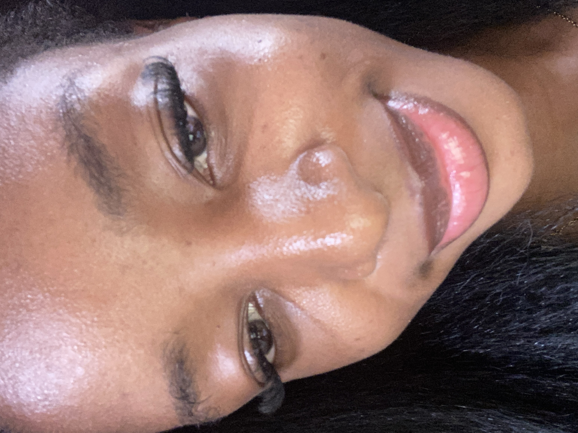 A close-up, low-angle, eye-level shot shows the face of a Black woman in Jos, Nigeria. Her dark, curly hair frames the left side of her face, and she is looking slightly downward and to her right. Her makeup includes voluminous false eyelashes, a subtle eyeshadow, and glossy pink lipstick. Her skin appears dewy, with highlights on her forehead, nose, and cheekbones. The background is dark and out of focus.