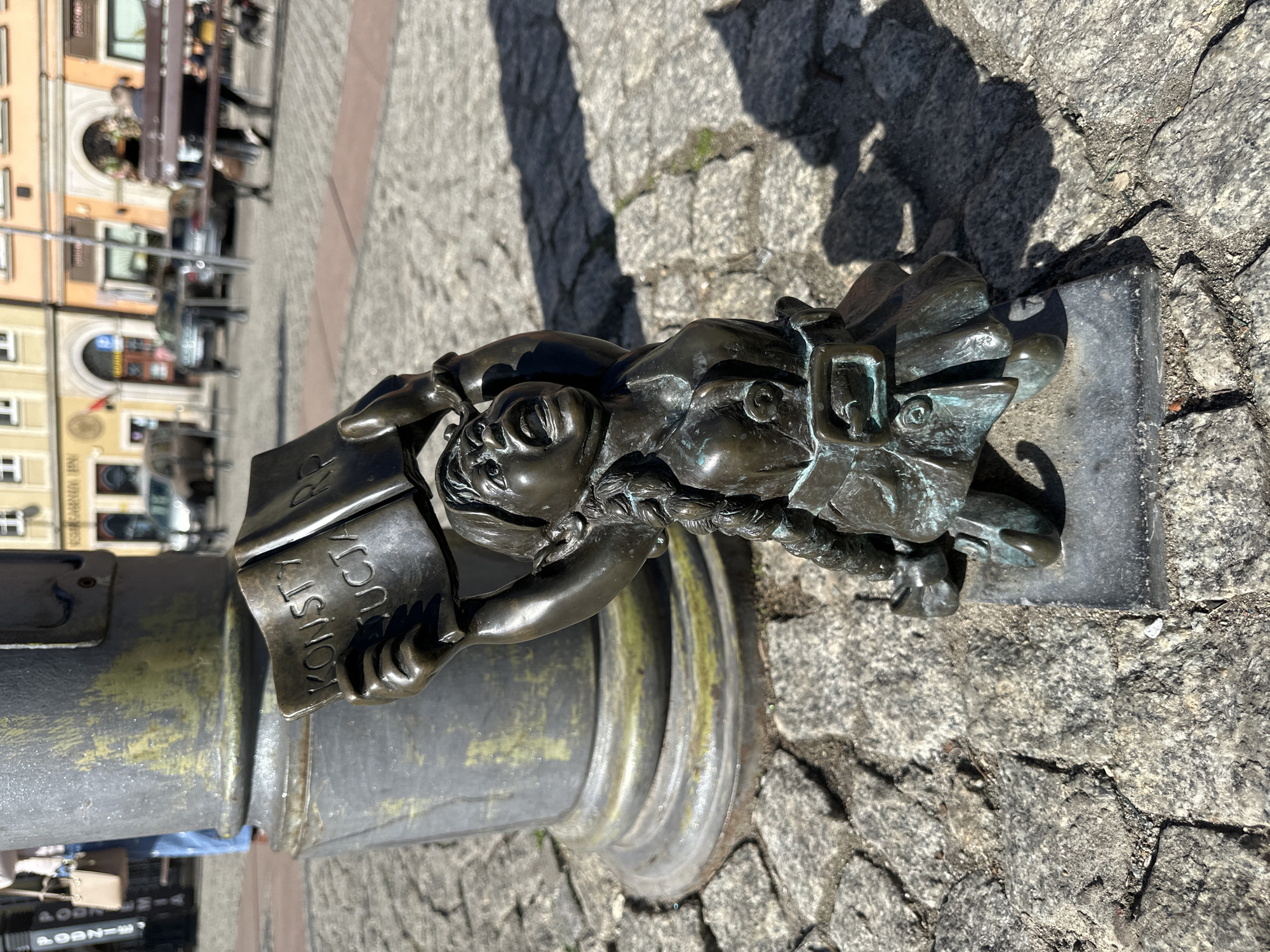 This image shows a bronze dwarf statue in Wrocław, Poland. The dwarf is attached to a metal post and is holding a book that reads "KONSTYTUCJA RP". The statue is located on a cobblestone surface in an outdoor area with buildings and cars visible in the blurred background. The scene is captured in daylight with shadows cast on the cobblestones.