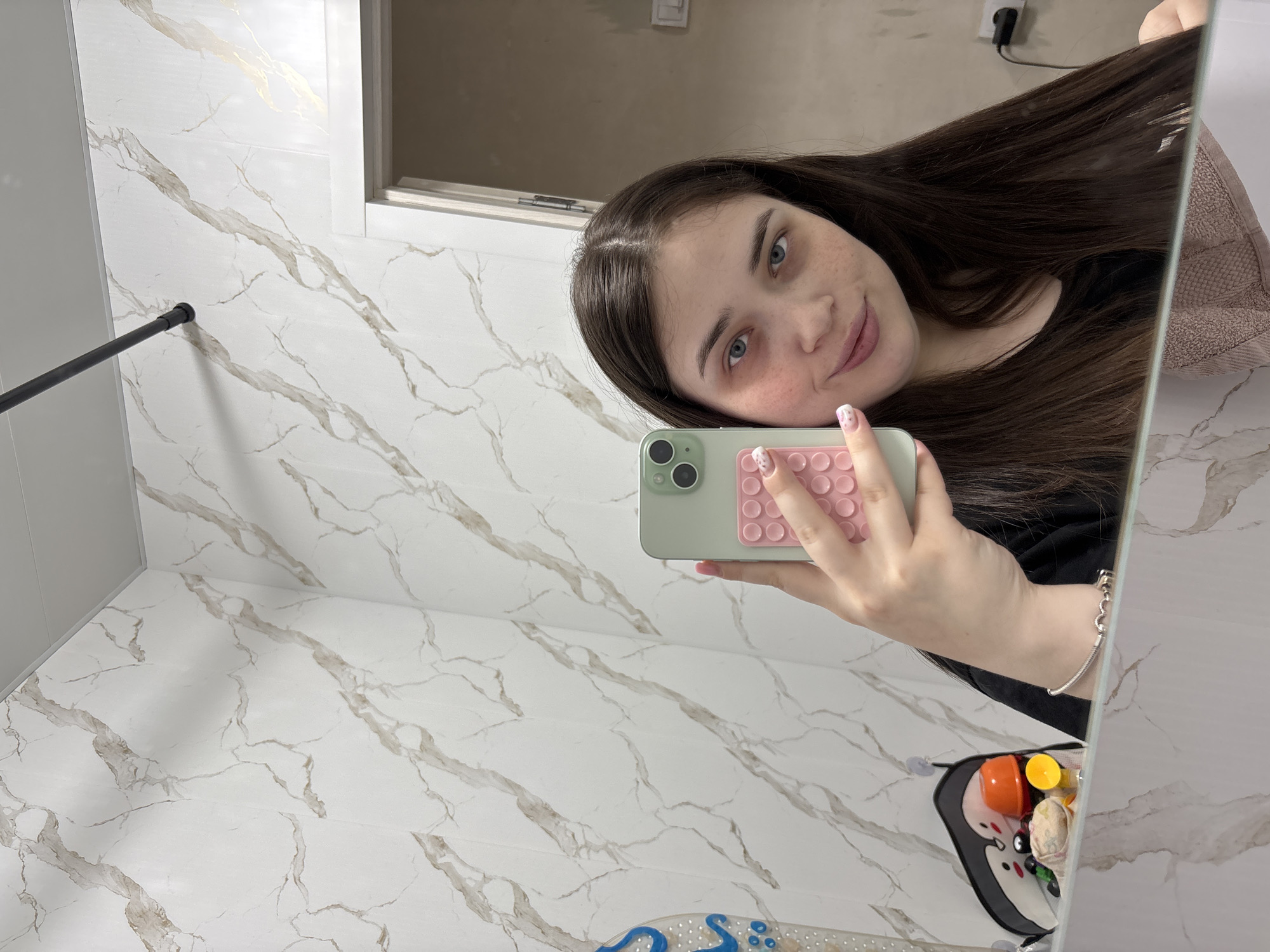 A woman is taking a selfie in a bathroom. She is holding a smartphone with a green case and a pink grip featuring circular patterns. The phone partially obscures her face. She has long, dark brown hair and light eyes. The background is a bathroom with marble-patterned tile on the walls and floor. A shower rod is visible on the left side of the frame. An electrical outlet is visible at the top of the frame. A toy organizer is visible at the bottom of the frame. The location is Chaikovsky, Russia.