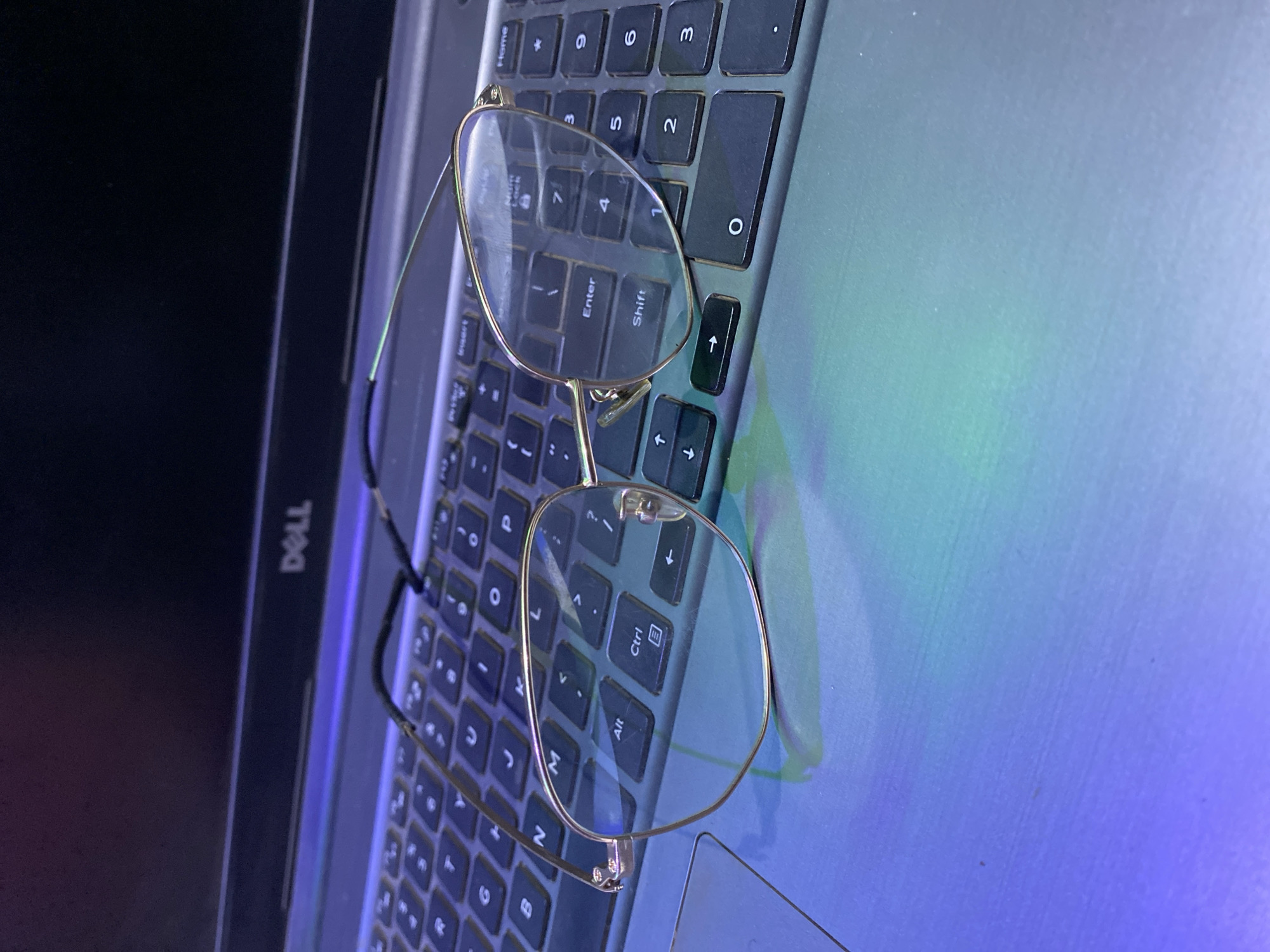 The image shows a pair of eyeglasses resting on the keyboard of a Dell laptop. The eyeglasses have thin, gold-colored frames and clear lenses. The laptop keyboard is black and grey, and the laptop lid is partially open. There is a screen to the left that is off. The background appears to be illuminated with blue and green light. There is no indication of the location of Bukuru, Nigeria, or any other element pertaining to that location.