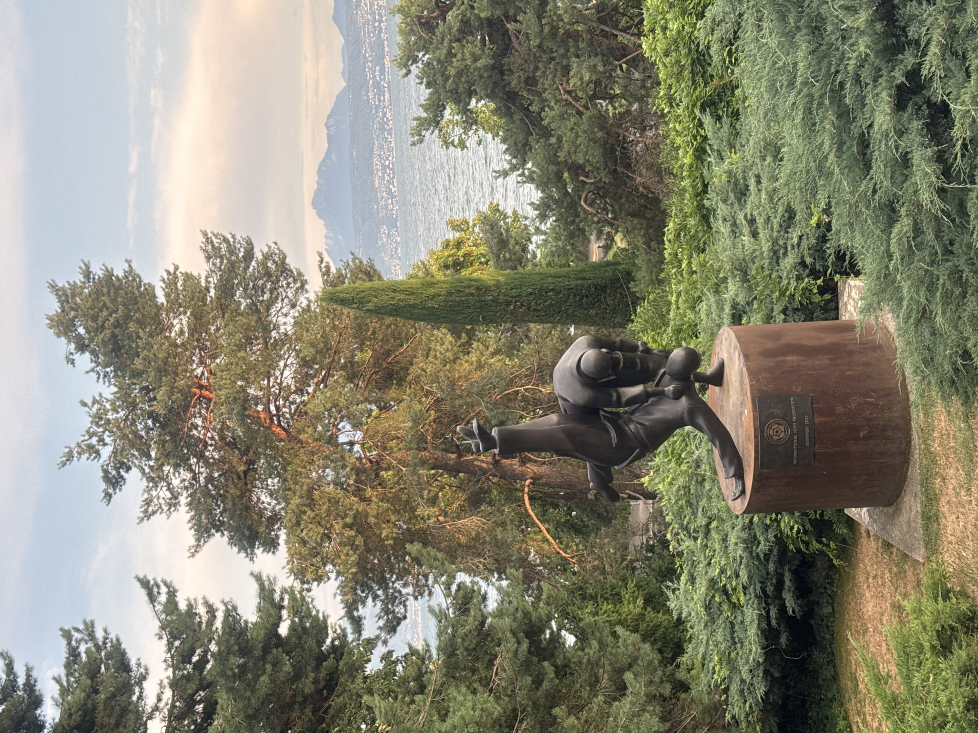 The image depicts a bronze sculpture of two judokas in combat, positioned on a cylindrical pedestal. The sculpture is set within a lush, green garden with dense shrubs and trees surrounding it. In the background, a body of water, likely Lake Geneva, is visible, with distant mountains under a cloudy sky. The pedestal of the sculpture features a plaque inscribed with "INTERNATIONAL JUDO FEDERATION BUDAPEST 2017". The scene appears to be in a park or outdoor public space, suggesting a location in Lausanne, Switzerland, known for its beautiful lakeside setting and numerous green areas. The lighting suggests it is either early morning or late afternoon.