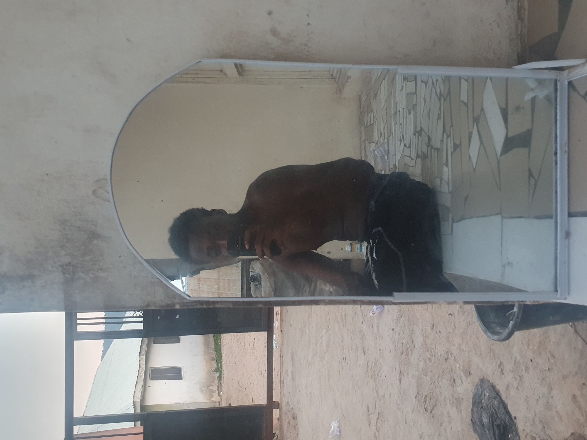 The image, rotated 90 degrees clockwise from its correct orientation, depicts a shirtless dark-skinned man taking a selfie in an upright mirror outdoors. The man, visible from the chest up, holds a dark smartphone in his right hand, aiming it towards his reflection. He wears dark-colored bottoms.
The mirror features an arched top and a white frame. Within the reflection, a background of light-colored walls is visible, one of which displays an irregular, geometric pattern of tiles in shades of white, grey, and beige.
Outside the mirror's frame, to the left, there's a light-colored, stained wall. The ground beneath the mirror is sandy or dusty earth, strewn with scattered white plastic debris, a dark bucket, and another dark, unidentifiable object.
In the background, beyond a dark metal gate or railing, are simple residential buildings. One structure has a light-colored corrugated roof and walls, featuring two small, dark-paned windows. Patches of green vegetation are also visible.
The scene is lit by natural daylight, indicating it is daytime. The setting appears to be a casual outdoor or semi-outdoor space, possibly a yard or open area, fitting for the location context of Lokoja, Nigeria. The man's expression is neutral, focused on his activity. There is no visible text in the image.