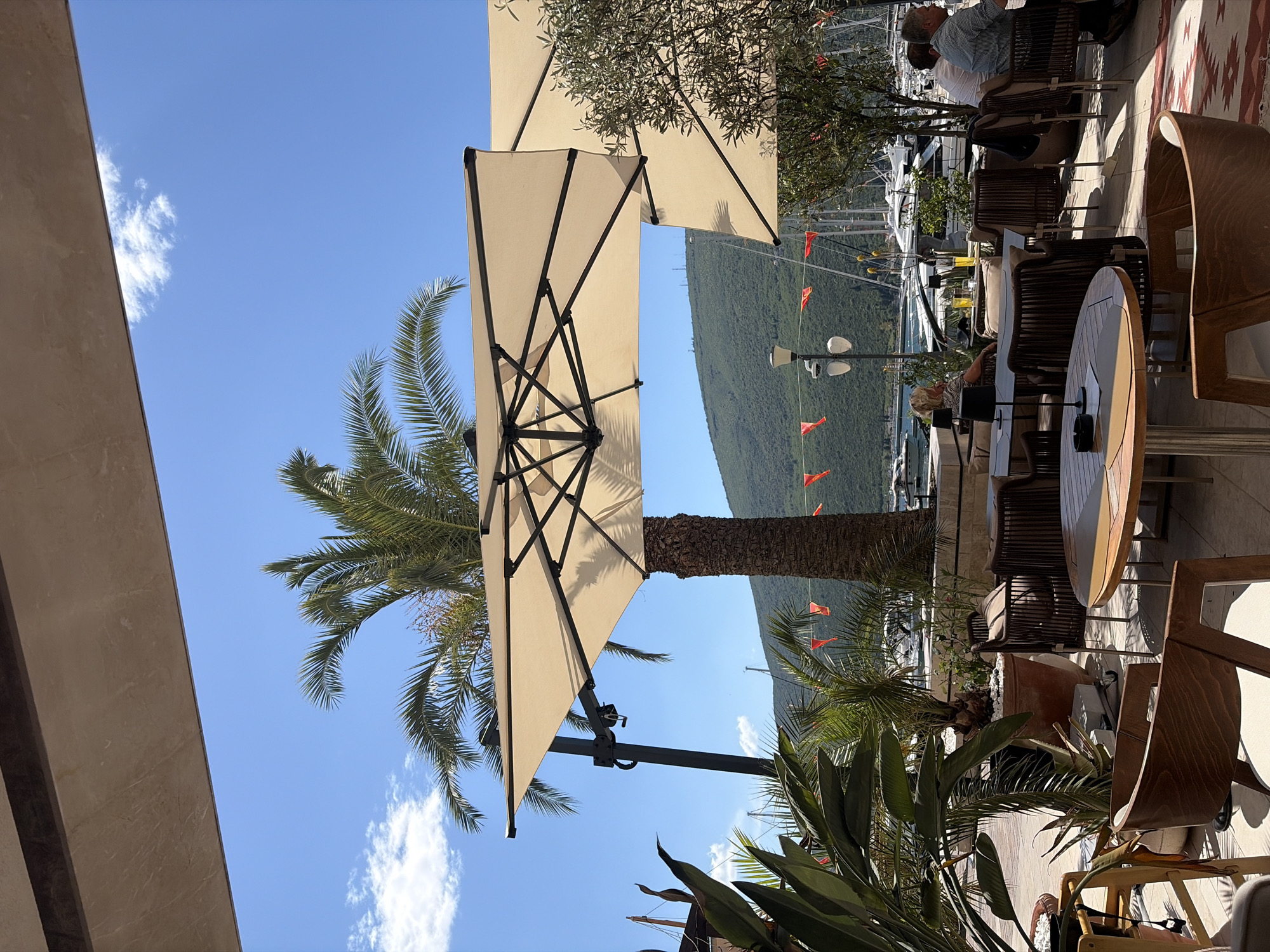 The image shows an outdoor café scene in Đenovići, Montenegro. The focus is on a partially opened cream-colored parasol supported by a central pole, casting shade over the tables and chairs. To the left of the parasol, a palm tree with green fronds stands against a clear blue sky with scattered white clouds. In the background, a hillside covered in green vegetation rises above a marina filled with boats. Orange flags are strung along the marina. The café area features wooden chairs, round tables, and various potted plants, including tropical foliage. People are seated at some of the tables, their actions and interactions partially obscured by the tables and parasol. The foreground includes architectural details of the café structure.