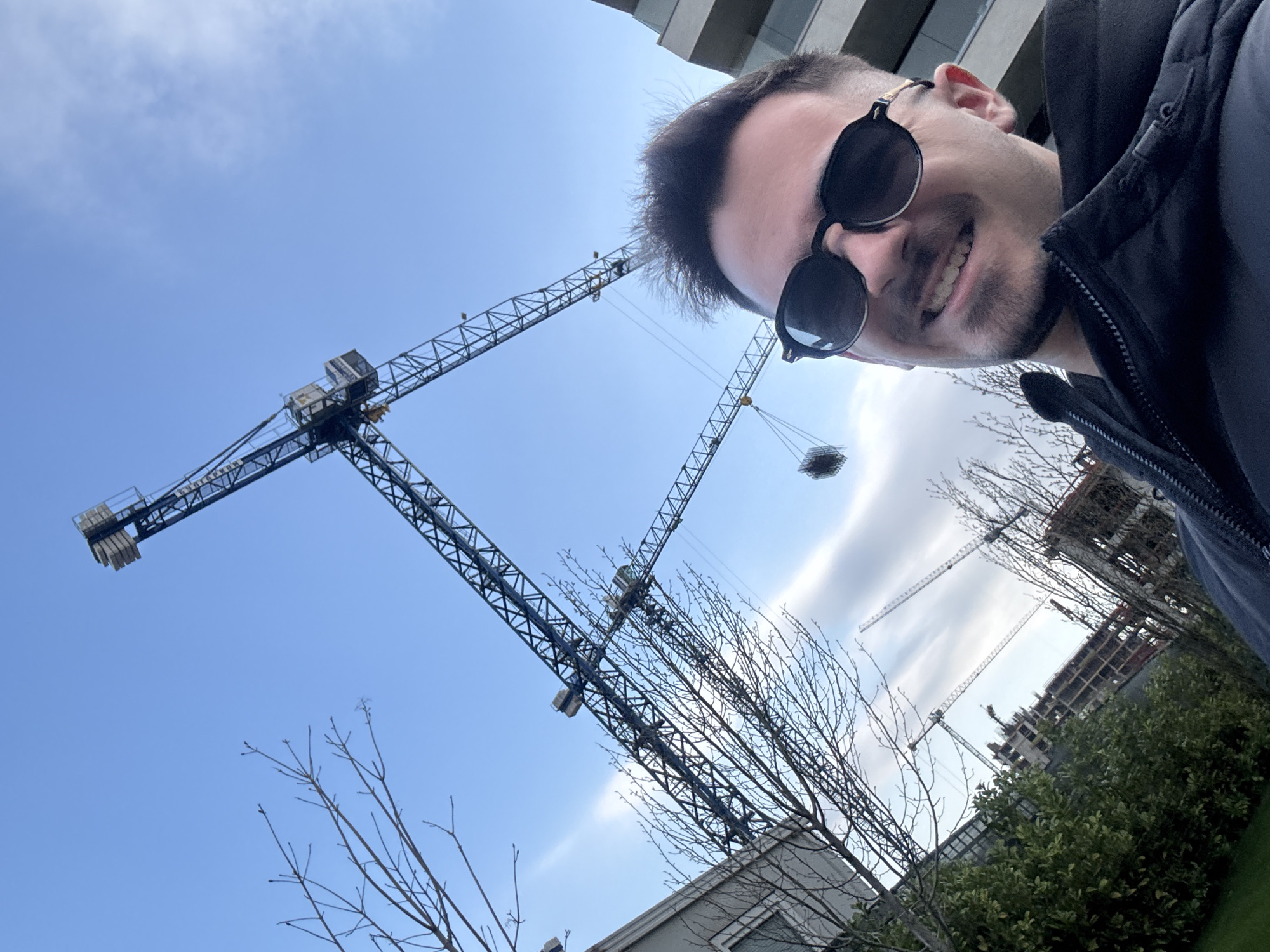 A man wearing sunglasses and a dark jacket is taking a selfie. Behind him, in a low-angle shot, are several construction cranes against a blue sky with some clouds. Bare trees are visible between the cranes. In the background, a building under construction is also visible. It appears to be a construction site in an urban environment. The construction site is in the area of Sofia, Bulgaria.