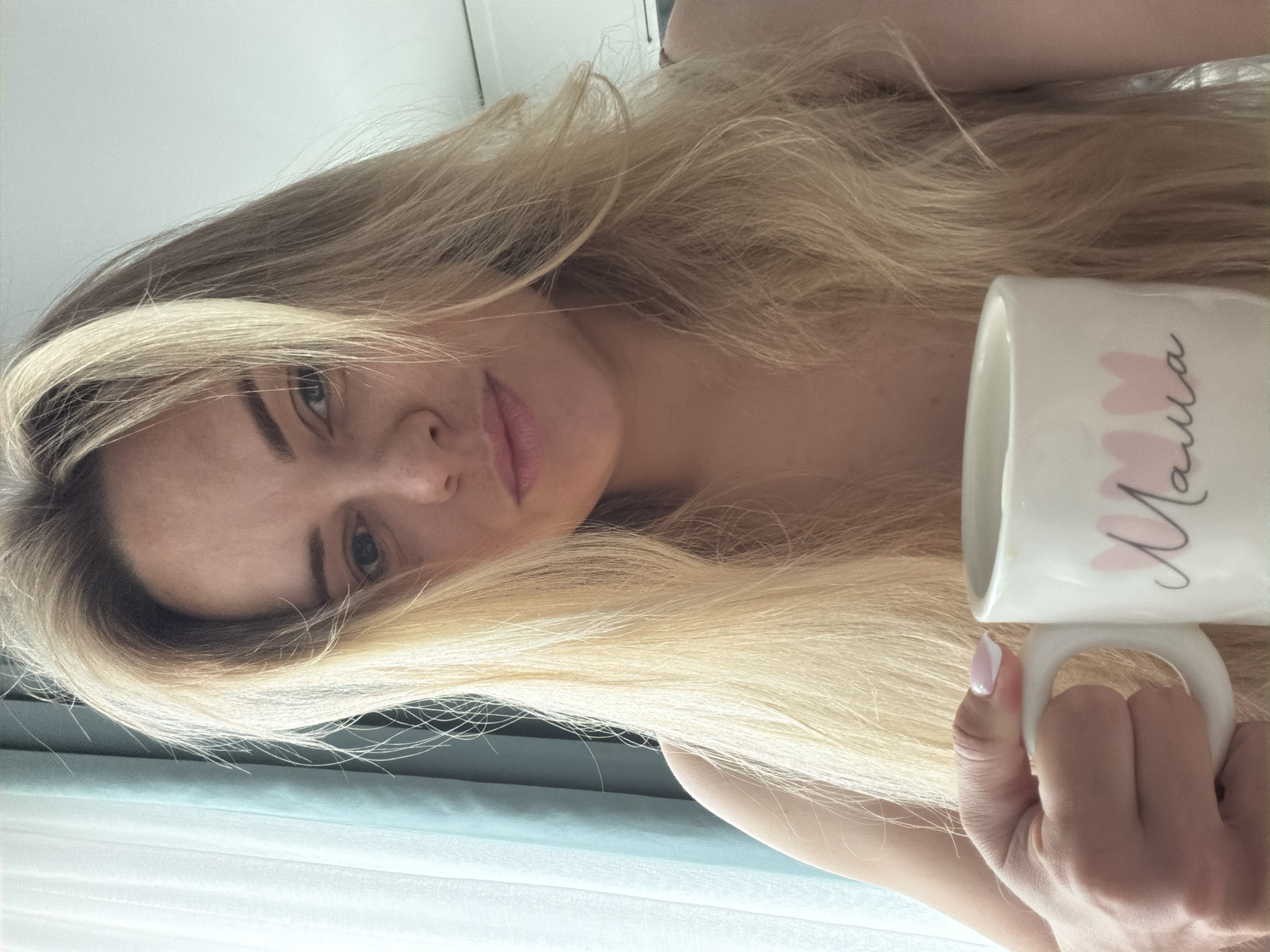A woman with long, light blond hair is holding a white mug with the word "Mama" written on it in cursive with pink heart accents. Her hand, with a painted nail, grips the mug's handle. The background appears to be an interior setting near a window with sheer curtains. It is not possible to determine the location in Votkinsk, Russia, from this image.