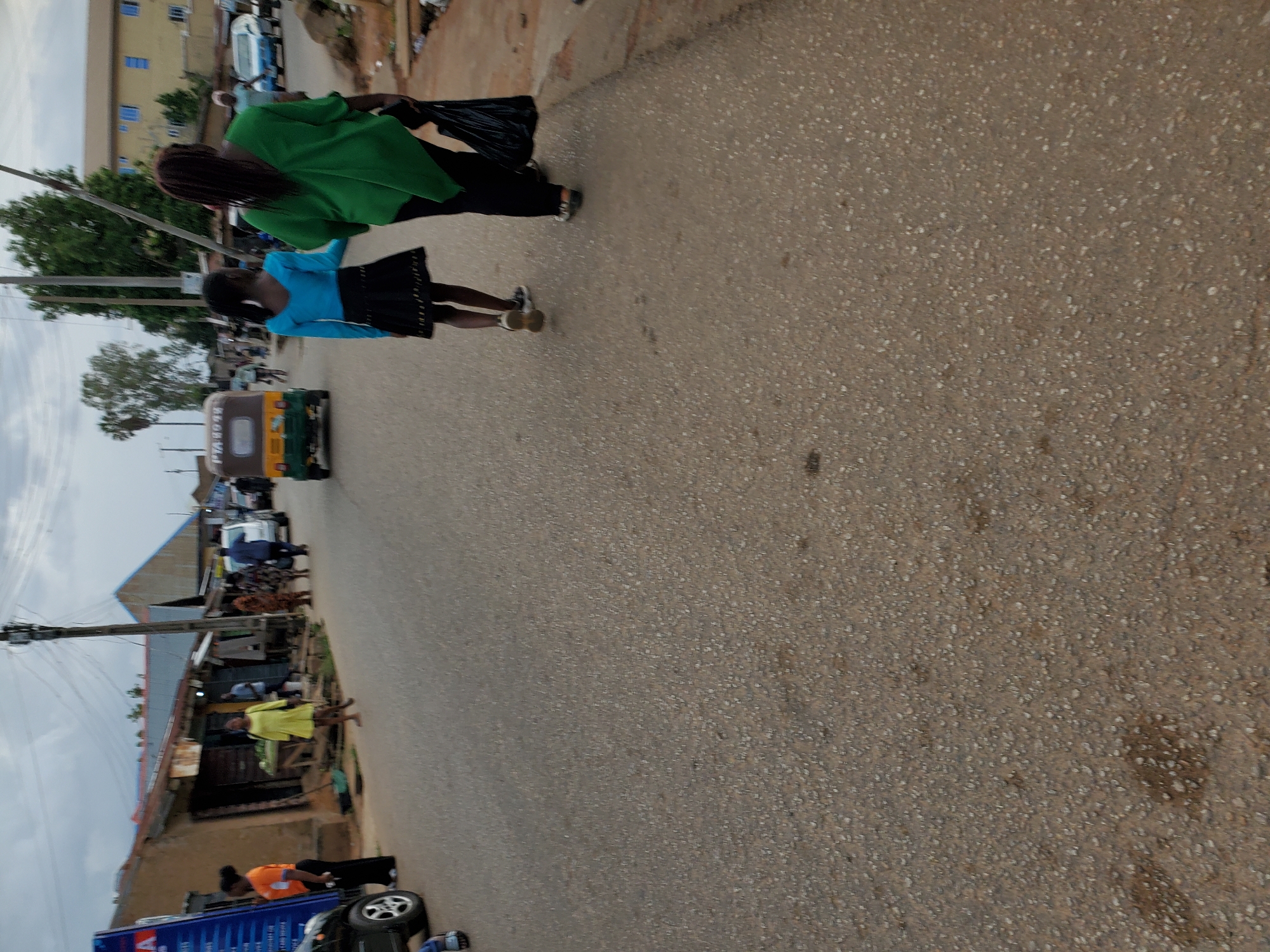 The image depicts a street scene in Jos, Nigeria. Two people, an adult wearing a green top and a child wearing a blue top and black skirt, are walking away from the camera on the street. A three-wheeled vehicle (a keke or auto-rickshaw) is traveling in the same direction, further down the road. Shops are lining the sides of the street, with people standing and sitting outside. There is a car parked on the left side of the road. Other vehicles and buildings are visible in the background. The road itself is paved. The sky is overcast.