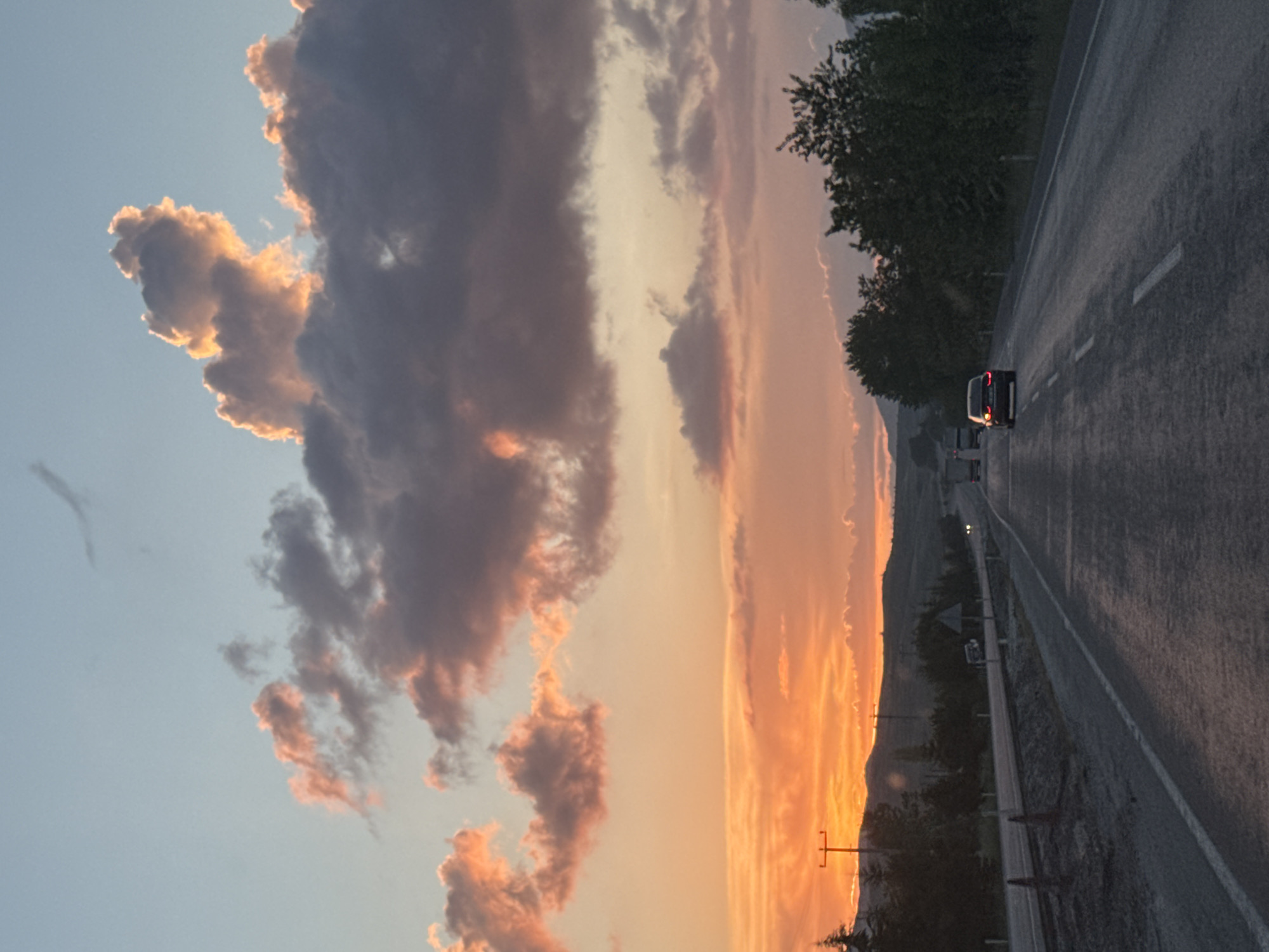 This image depicts a rural highway scene in Korkuteli, Turkey, at dusk. The sky is characterized by a vibrant sunset, displaying a gradient from a pale blue at the top to a dominant orange and reddish-pink hue along the horizon. Cumulus clouds are present across the sky, with some illuminated in bright orange and yellow by the setting sun on the upper right, while others appear as darker grey or reddish-purple formations.
The foreground is dominated by a two-lane asphalt road that recedes into the distance. The road surface appears damp, reflecting the ambient light. White dashed lines delineate the lanes, and solid white lines mark the shoulders. On the left side of the road, a dirt shoulder is visible, alongside what appear to be traffic cones or temporary barriers. A dark coniferous tree and two utility poles with overhead lines are situated on the left, silhouetted against the bright sky.
In the mid-ground, a dark sedan with illuminated taillights is present in the right lane, moving away from the viewer. Further down the road, another vehicle with illuminated headlights is visible in the same lane, and multiple trucks or vehicles with headlights are discernible in the opposing lane on the far left. A white vehicle is parked or stopped on the left shoulder further into the distance, near a triangular road sign. The surrounding environment consists of dark green trees and bushes lining both sides of the road, backed by silhouetted hills or mountains along the horizon.