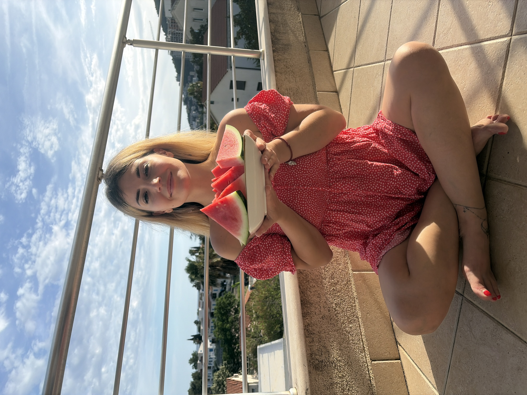 A young adult female sits cross-legged on a light-colored tiled balcony floor in Budva, Montenegro, under a blue sky with scattered white clouds. She has long blonde hair and wears a red off-the-shoulder dress with small white polka dots. She smiles faintly while looking directly at the camera. She holds a white, oval-shaped plate in front of her with both hands, presenting multiple triangular slices of red watermelon. Her feet are bare, with red nail polish on her toenails, and a delicate linear tattoo with a crescent moon design is visible on her left ankle. She wears a dangle earring in her visible right ear and a thin red bracelet on her left wrist. The balcony features a textured, light brown knee-wall and a silver-colored metal railing with multiple horizontal bars. The background reveals a coastal city scene with numerous white and light-colored buildings featuring red-tiled roofs, interspersed with green trees and hills. A construction crane is visible on the right side of the distant cityscape. Strong sunlight creates distinct shadows on the balcony surfaces.