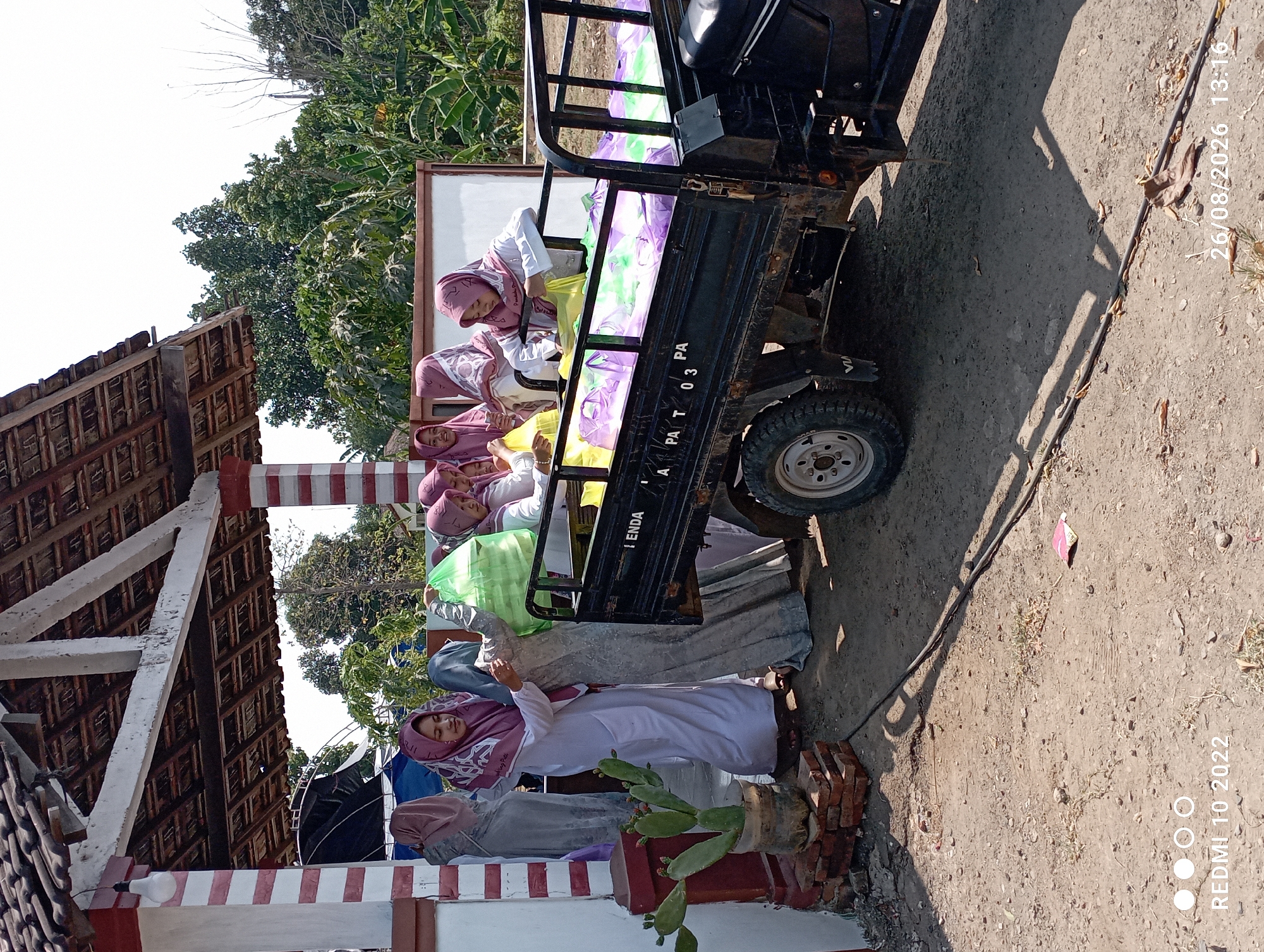 In Kecamatan Pati, Indonesia, a group of women wearing hijabs are engaged in loading bags into the back of a small truck. The truck is black and appears to be a utility vehicle. The bags are predominantly green, purple, and yellow, suggesting they contain some sort of goods or supplies. The women are standing and interacting with each other as they transfer the bags. One woman is actively lifting a green bag, while others appear to be organizing or placing bags into the truck.
The setting appears to be outdoors, with a building visible in the background. The building has a tiled roof and white-painted supports. There are trees and foliage surrounding the area, indicating a rural or semi-rural environment. A cactus plant in a pot is situated near the building, adding to the outdoor scene. The ground is dusty and appears to be unpaved. The lighting suggests it is a sunny day. The timestamp "REDMI 10 2022" and "26/08/2026 13:16" are visible in the lower portion of the image.