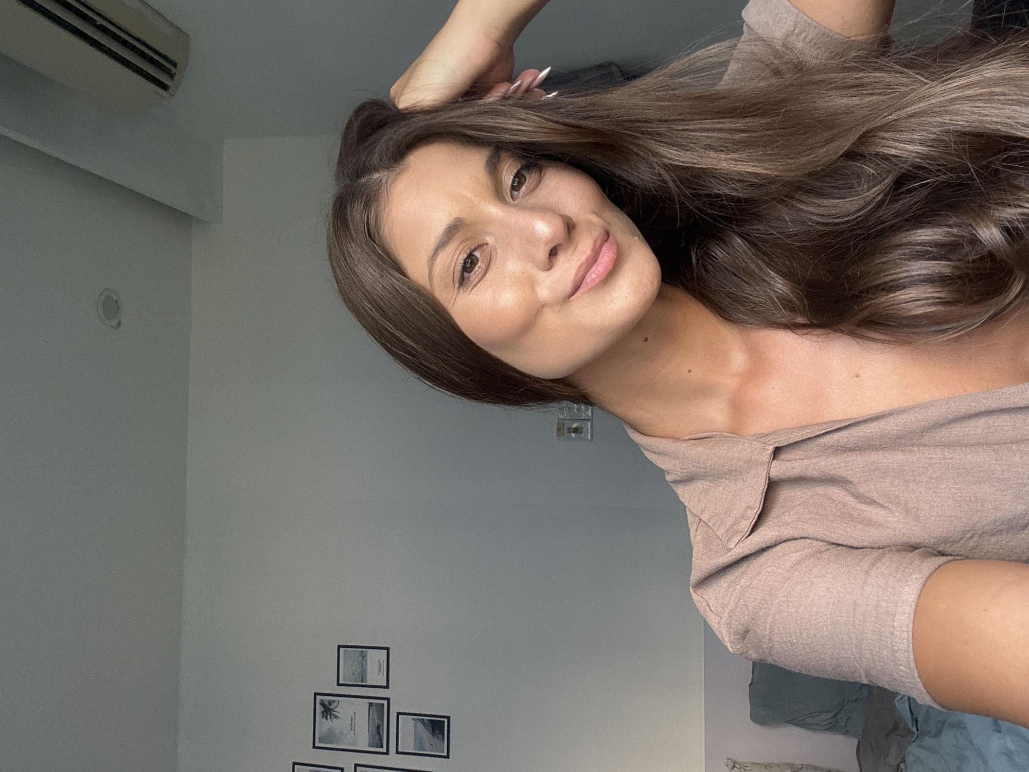 A woman with long, brown hair is posing for a photo indoors. She is holding her hair with her right hand and smiling slightly. She is wearing a light brown top with a collar. The background is a light gray wall with a few framed pictures on the lower left side and an air conditioning unit in the upper left corner. The location in the city of Đà Nẵng, Vietnam cannot be confirmed.