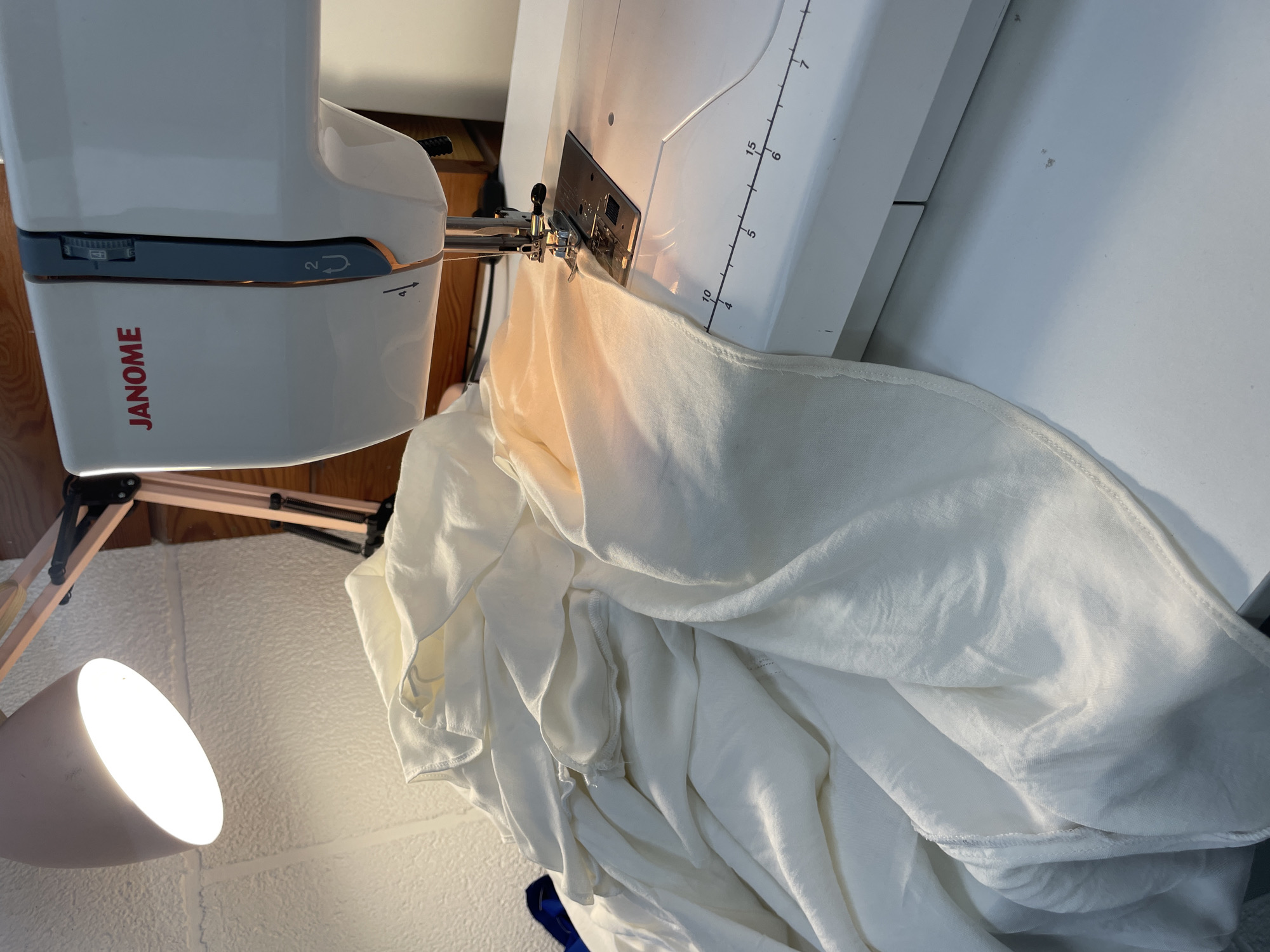 The image shows a close-up view of a Janome sewing machine with white fabric. The sewing machine is on a white table, and a task lamp is illuminating the fabric and the sewing machine. The fabric appears to be a light, flowing material, possibly silk or a similar synthetic. The sewing machine is in operation, with the needle positioned to stitch the fabric. The background consists of a textured wall and a wooden surface. The scene is indoor and well-lit, suggesting a workspace or craft area. There are no people visible in the image. The location in the city of Sinyavino, Russia, is not discernible from the visual information provided in the image.