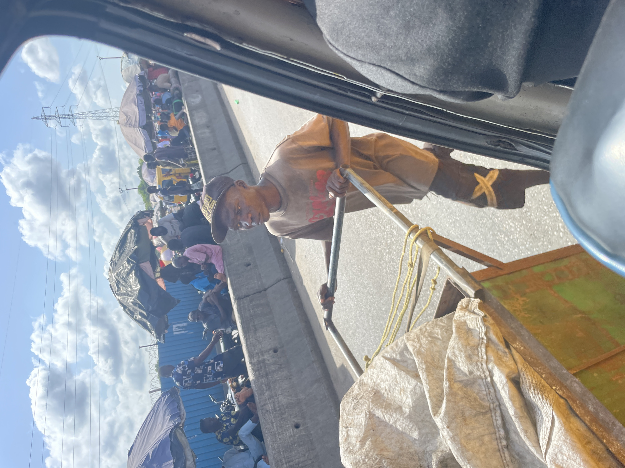 The image, taken from inside a vehicle looking out a window, captures a vibrant street scene in Alasia, Nigeria, under bright daylight with scattered white clouds in a blue sky.
In the immediate foreground, the interior of the vehicle is visible. Just outside, a man is seen pushing a metal cart along a paved surface, likely a road. He is wearing a light brown t-shirt, a dark cap, and dark boots, one of which features a distinctive light-colored "X" design. The man looks directly towards the camera with a neutral, perhaps focused, expression. His cart carries items, including a large white, possibly fabric, bag and yellow ropes, with a greenish-brown base visible.
A concrete barrier runs parallel to the road, separating the man from a densely populated area beyond. This area is bustling with numerous people, some standing, many gathered or seated under various makeshift shelters and umbrellas, suggesting a busy market or roadside vending area. Tarps and tents are used to create these temporary structures, indicating commercial activity.
In the background, beyond the crowded market area, a tall metal power pylon stands prominently against the sky. The overall scene depicts a dynamic outdoor environment, filled with human activity and daily commerce under clear, sunny conditions. No clearly legible text is visible on clothing or signs.