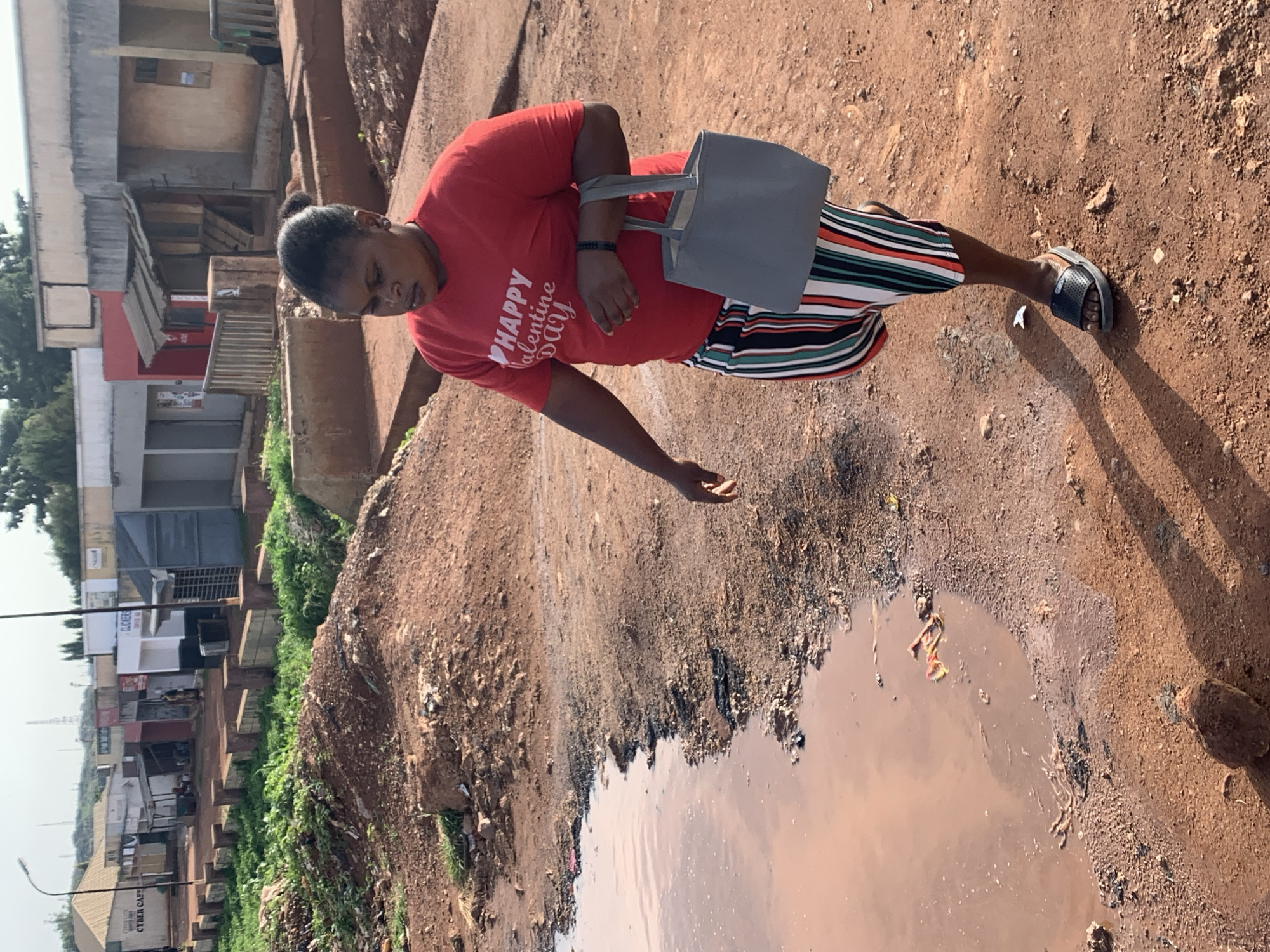 A woman walks on a dirt path next to a muddy puddle in Gura Top, Nigeria. She wears a red t-shirt with "Happy Valentine Day" written on it in white lettering, and striped pants in green, white, red, and black. She carries a gray tote bag in her right hand and wears black sandals. The environment is arid with red dirt, sparse green vegetation, and several low-lying buildings in the background.