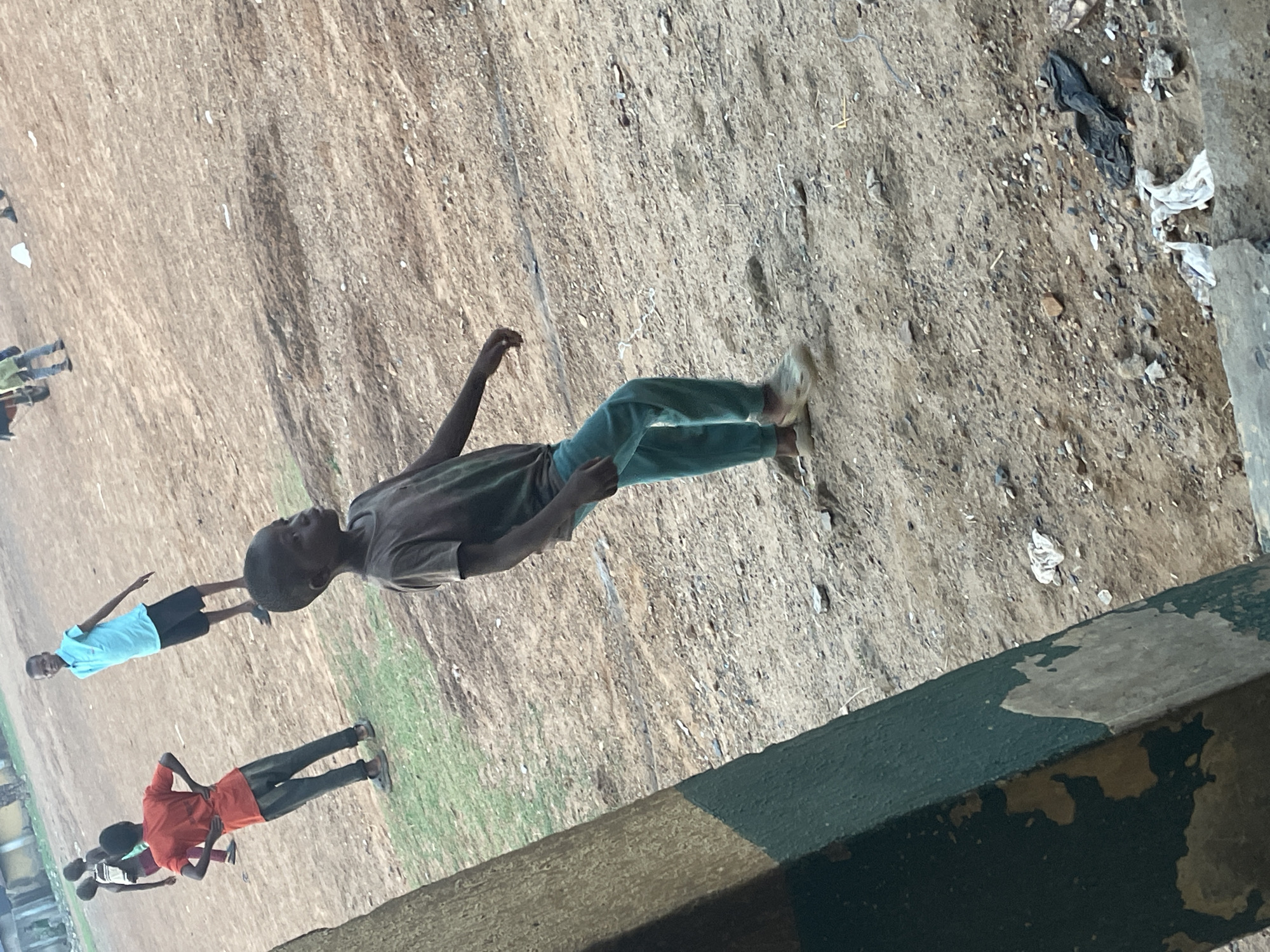 The image shows a dusty field on the outskirts of Akwanga, Nigeria. In the foreground is a green painted concrete barrier, likely a wall or the edge of a building. On the field, there are several young people, possibly boys, engaged in what appears to be a casual activity or game. One individual is closer to the viewer, wearing a brown shirt and green pants, with one arm raised. Further away, others are seen wearing various colored shirts (red, blue), with darker colored shorts or pants. The ground is mostly barren, with scattered debris.