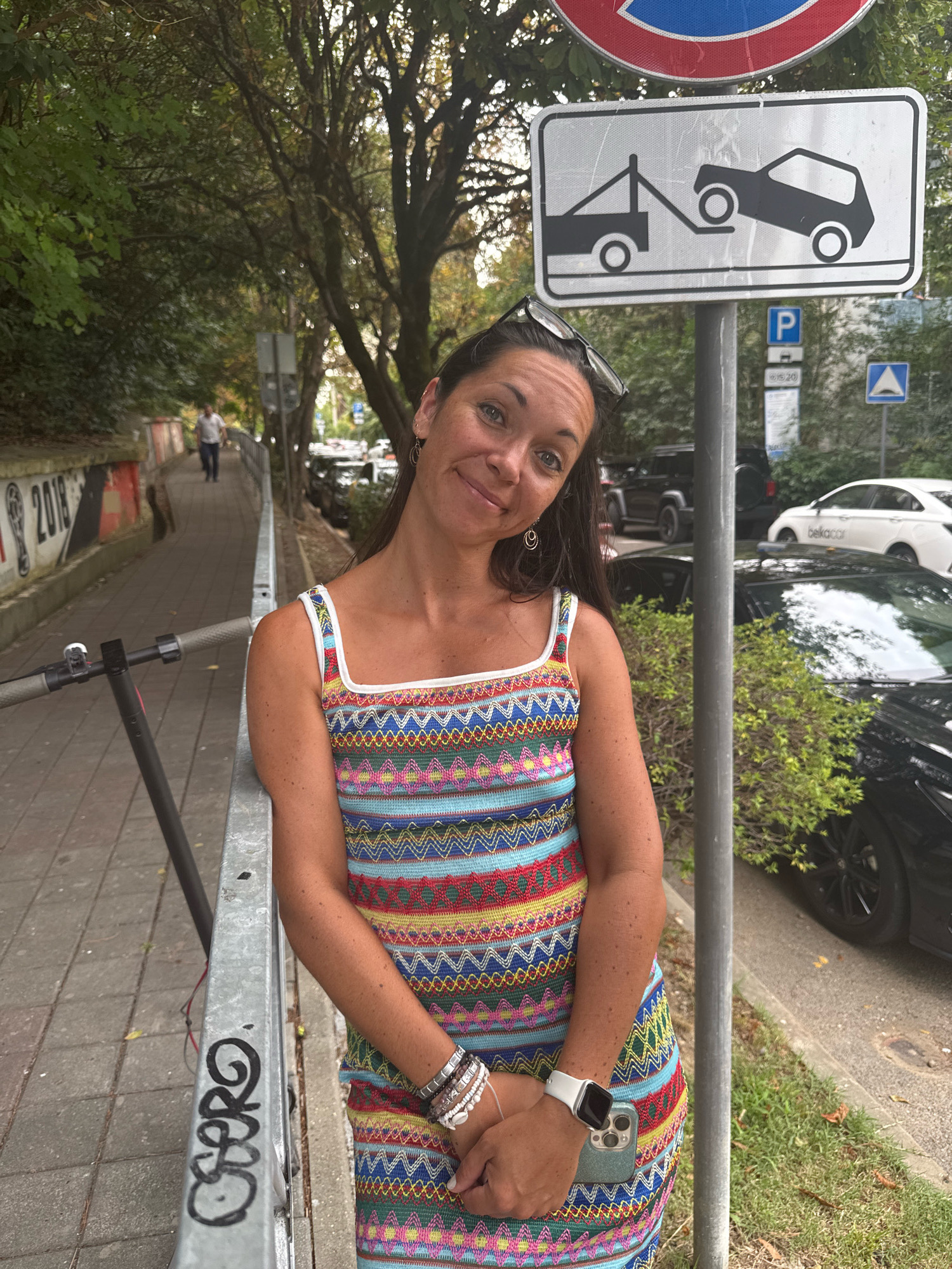 A medium shot captures a woman standing on a paved sidewalk next to a metal railing, in what appears to be a city park in Sochi, Russia. She is wearing a colorful, patterned dress and several bracelets on her left wrist. Her right hand holds a smartphone with a green case. Behind her, a towing sign is visible on a pole, indicating that vehicles will be towed. The background shows trees, parked cars, and a street with more trees. The overall scene depicts an outdoor urban environment, possibly a pedestrian walkway with adjacent parking.