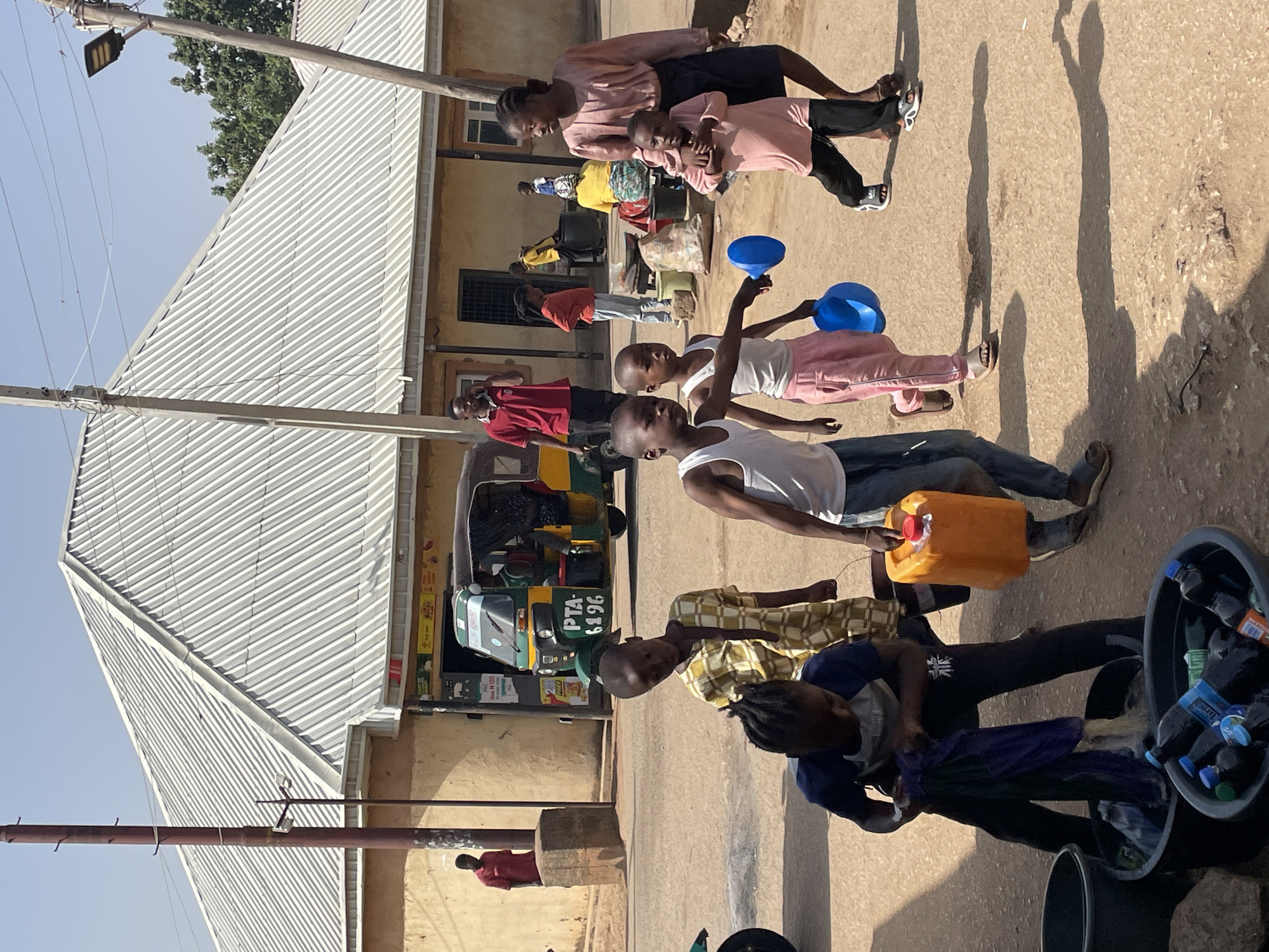 This daytime image, taken in Jos, Nigeria, captures a bustling street scene featuring numerous people, mainly children and young adults, engaged in various activities. The setting includes an unpaved road, basic buildings with corrugated metal roofs, and overhead utility lines.
In the foreground, several children are prominent. One child, wearing a white tank top and dark trousers, holds an orange jerrycan. Another child, dressed in a patterned yellow and white shirt, stands close by. To their right, a child in a blue and white shirt is attending to a black plastic tub filled with an assortment of plastic bottles, suggesting a commercial or collection activity. Further back, two children in white tank tops and light-colored pants are holding blue funnels or bowls. An adult woman in a pink top and dark skirt is seen with a younger child, also in pink.
A green and yellow tricycle, identified by the text "PTA 6196" on its side, is parked in the mid-ground. Other individuals are visible in the background, including someone in a red shirt standing near a building. The bright sun casts long, distinct shadows across the ground, indicating either morning or late afternoon. The sky is clear, and the scene depicts typical daily life in the community, with people dressed in casual clothing.