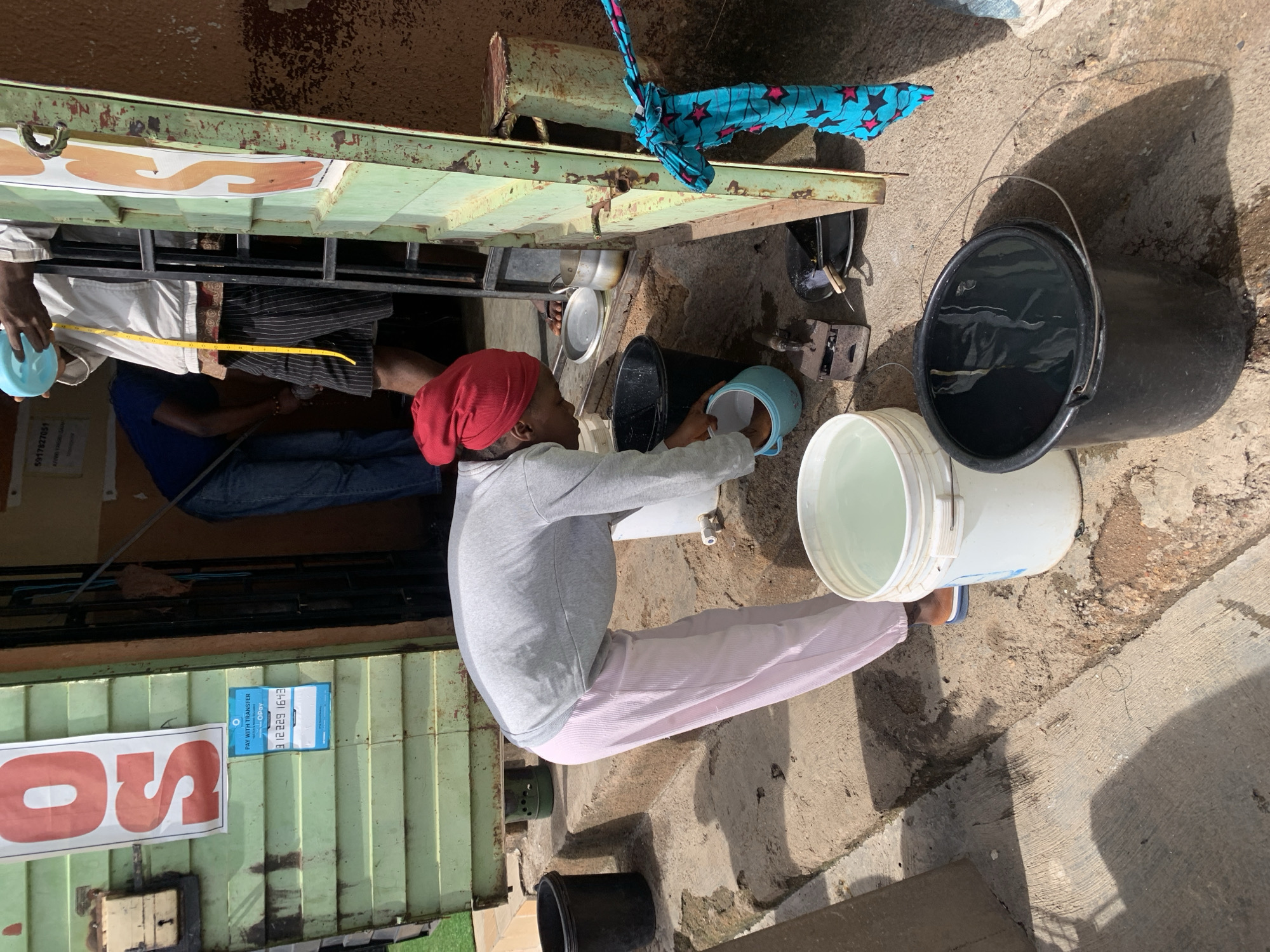 This image captures an outdoor scene, likely a water vending point or a small shop, in Jos, Nigeria, under bright daylight. The ground is paved with concrete, and strong shadows indicate a sunny day, possibly midday or early afternoon.
In the foreground, a person, potentially a woman or girl, is squatting low while engaged in an activity involving water. She wears a grey long-sleeved top, light pink striped pants, and a red head covering. She is holding a small light blue plastic bowl, seemingly filling it or transferring water from a source on a green metal counter.
The counter is part of a rustic, green-painted metal stall or kiosk, showing signs of wear and rust. On the counter, several small metal pots or bowls are visible, and a bright blue fabric with pink stars is draped over it. Behind this counter, another person in a white shirt and dark pants is partially visible, holding a yellow measuring tape. The legs of a third person in blue jeans are seen further back.
On the ground around the counter, multiple plastic buckets are present: a large black one partially filled with water, and a white one nearly full of water. An old-fashioned weighing scale with iron weights sits near the black bucket.
Visible text includes a red sign on the green counter with the partial word "OS". A blue and white sign attached to the counter reads "PAY WITH TRANSFER", features an "OPay" logo, and displays the phone number "91 2229 1642", with "POS" partly visible below it, suggesting mobile payment options. The scene depicts a common daily activity, likely the sale or distribution of water, set against the backdrop of a local commercial establishment.