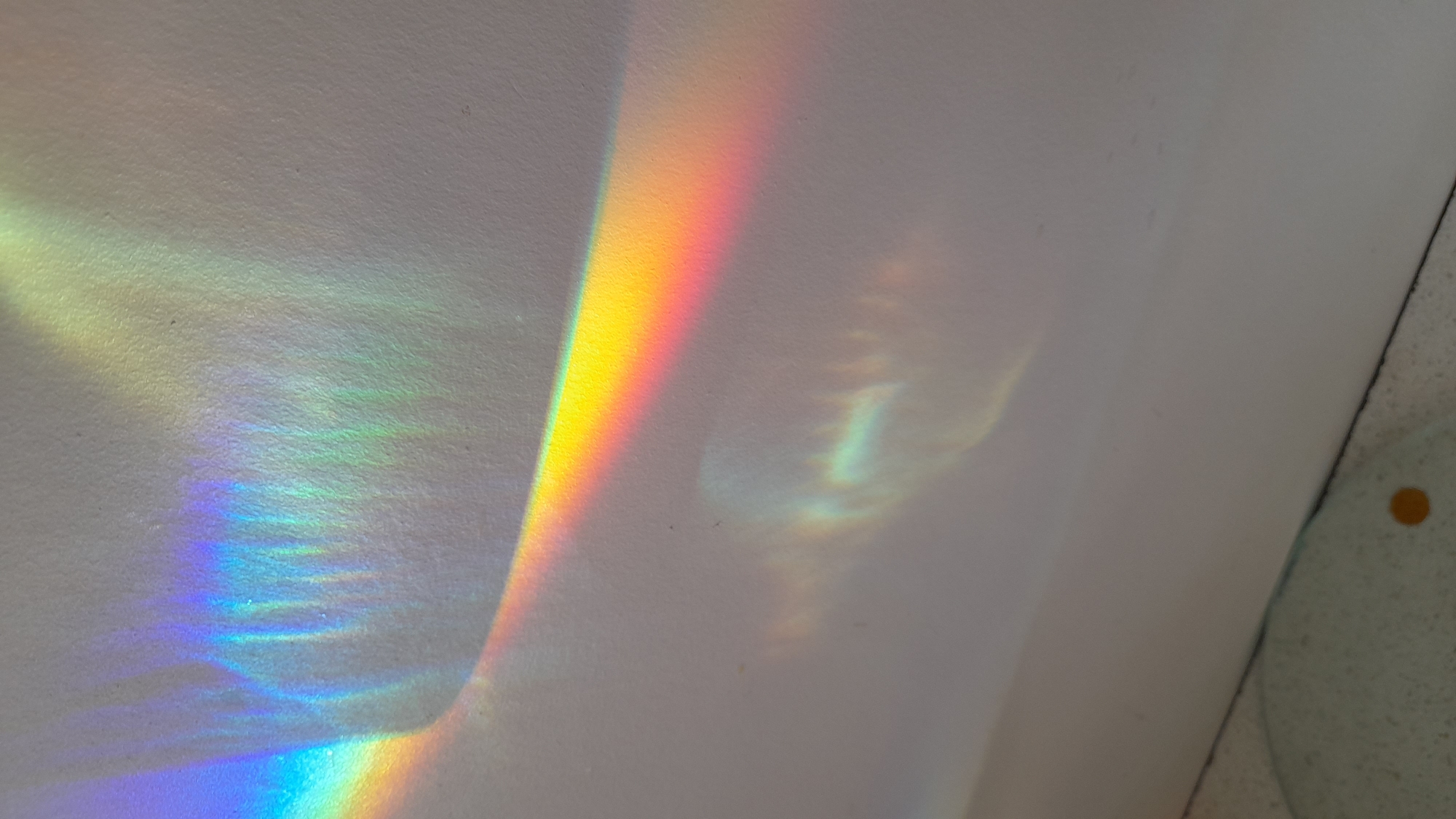 The image shows a section of a white wall illuminated by a rainbow spectrum of light. The colors are distinct, transitioning from blue and violet to green, yellow, and red. A secondary, fainter rainbow pattern is visible next to the brighter one, appearing as a ghost-like outline. Part of a light blue colored item with a single orange colored dot is visible in the lower right of the frame. The scene is indoor. There is no indication of the location being Saint Petersburg, Russia or any other city.