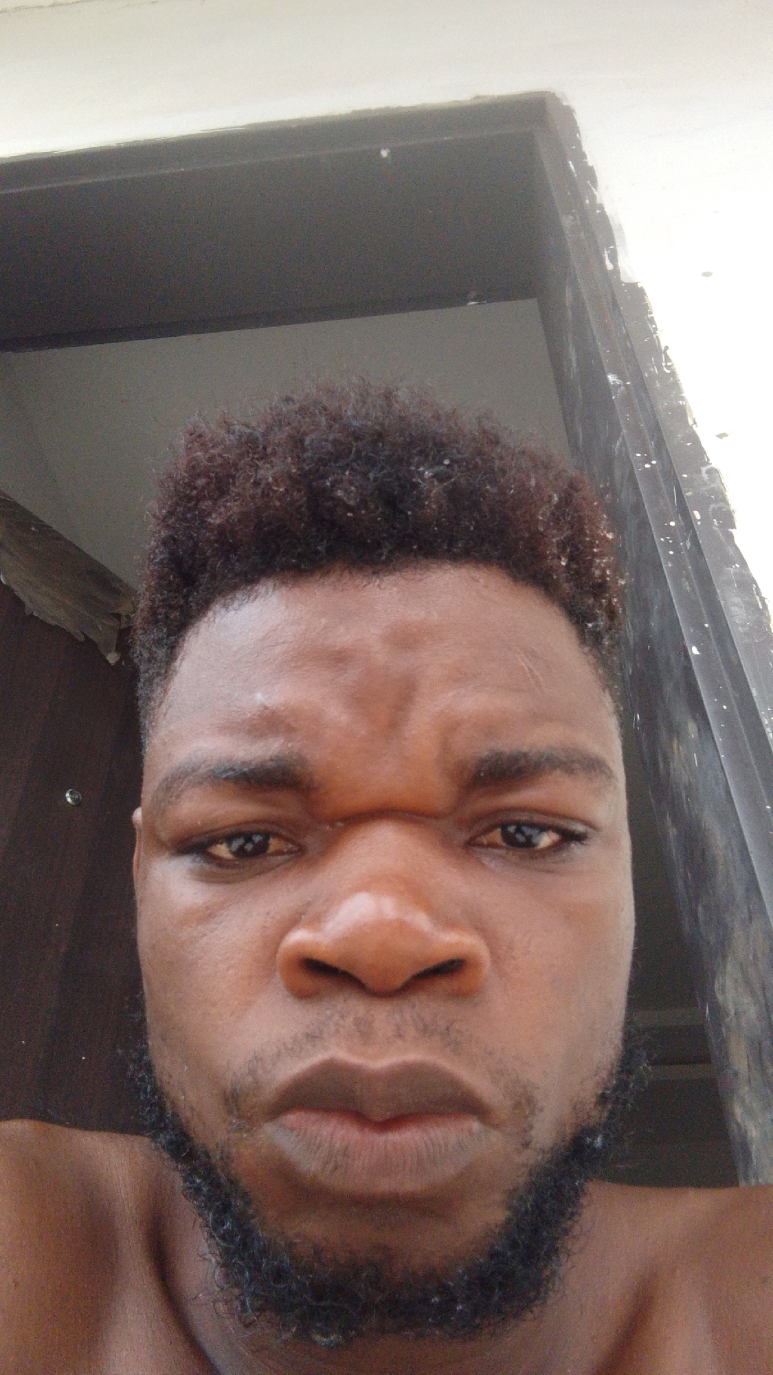 A close-up, front-facing selfie features an adult male with dark skin, likely taken in Enugwu-Ukwu, Nigeria. He is centered in the frame, looking directly into the camera with a serious or stern expression, characterized by furrowed brows and a direct gaze. He has dark, tightly curled hair and a full, short beard and mustache. His shoulders are visible, suggesting he is shirtless. The background consists of a dark door frame or entryway to the left and right, with a bright, light-colored ceiling or upper wall section visible above his head. The lighting appears to be natural, possibly from indoors or a shaded area. There is no visible text in the image.
