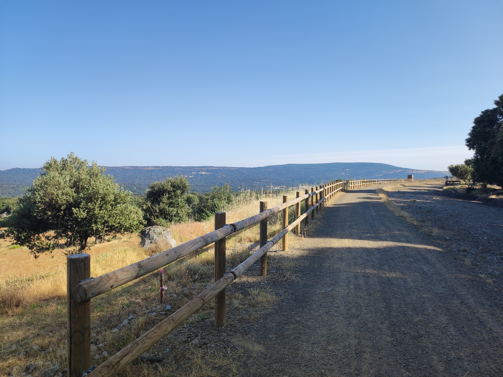 The image depicts a scenic rural landscape with a clear blue sky. A dirt path curves through the scene, bordered by a rustic wooden fence. The fence, constructed from rough-hewn logs, runs along the right side of the path and recedes into the distance. The terrain to the left of the path is covered in dry, yellow grass and scattered rocks, with a few trees present. In the background, a rolling, tree-covered hill creates a natural horizon. The lighting suggests it is either early morning or late afternoon, casting long shadows across the path. No people or animals are visible in the image. This scene is likely located in a rural area outside of Ledrada, Spain, given the characteristic Mediterranean vegetation and terrain.