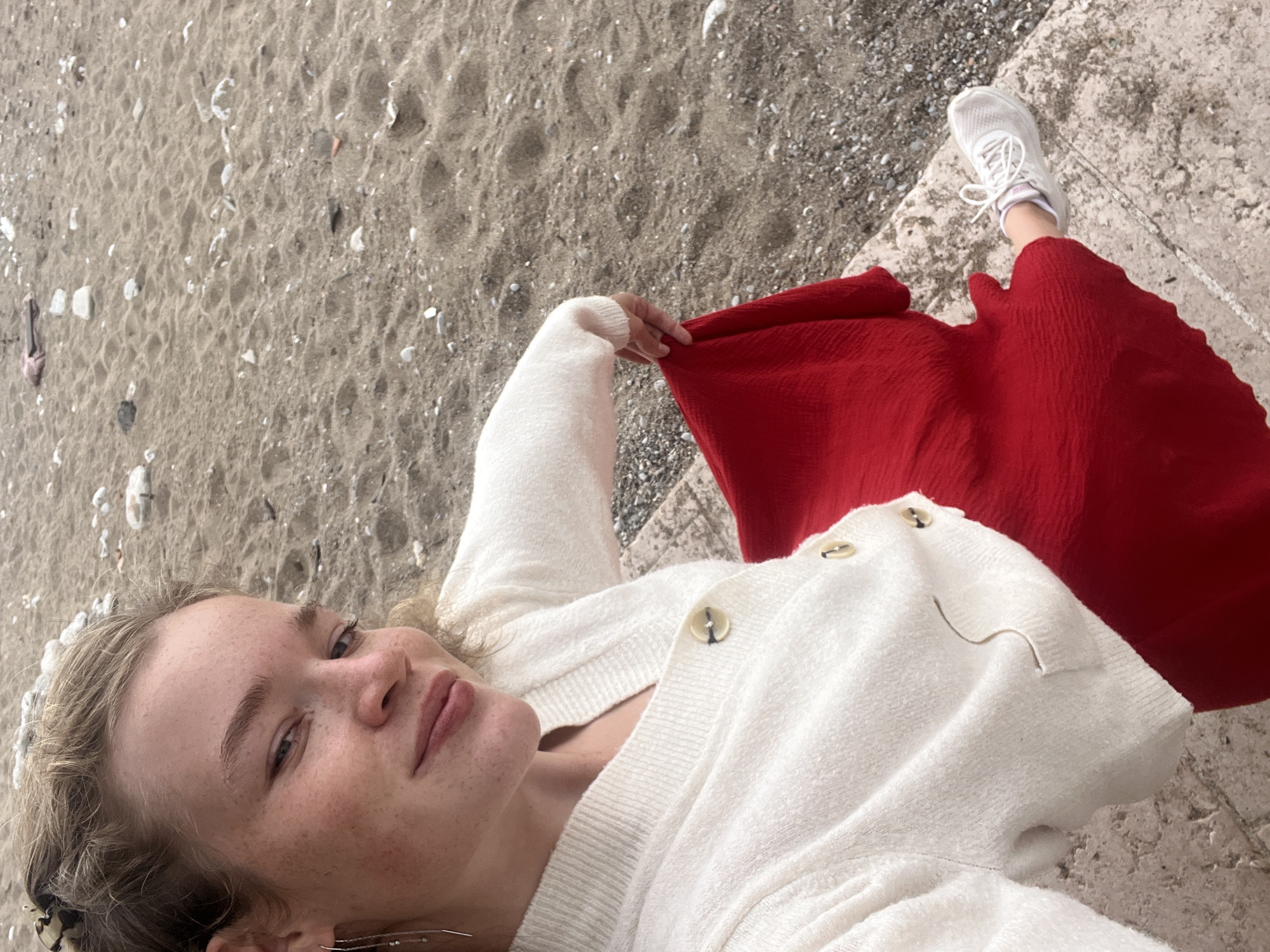 A woman with freckles is lying on the beach in Unknown, Unknown. She is wearing a white cardigan and a red dress. Her left leg and foot in a white sneaker are on a stone ledge. A beach of grey sand and white rocks lies to her left.