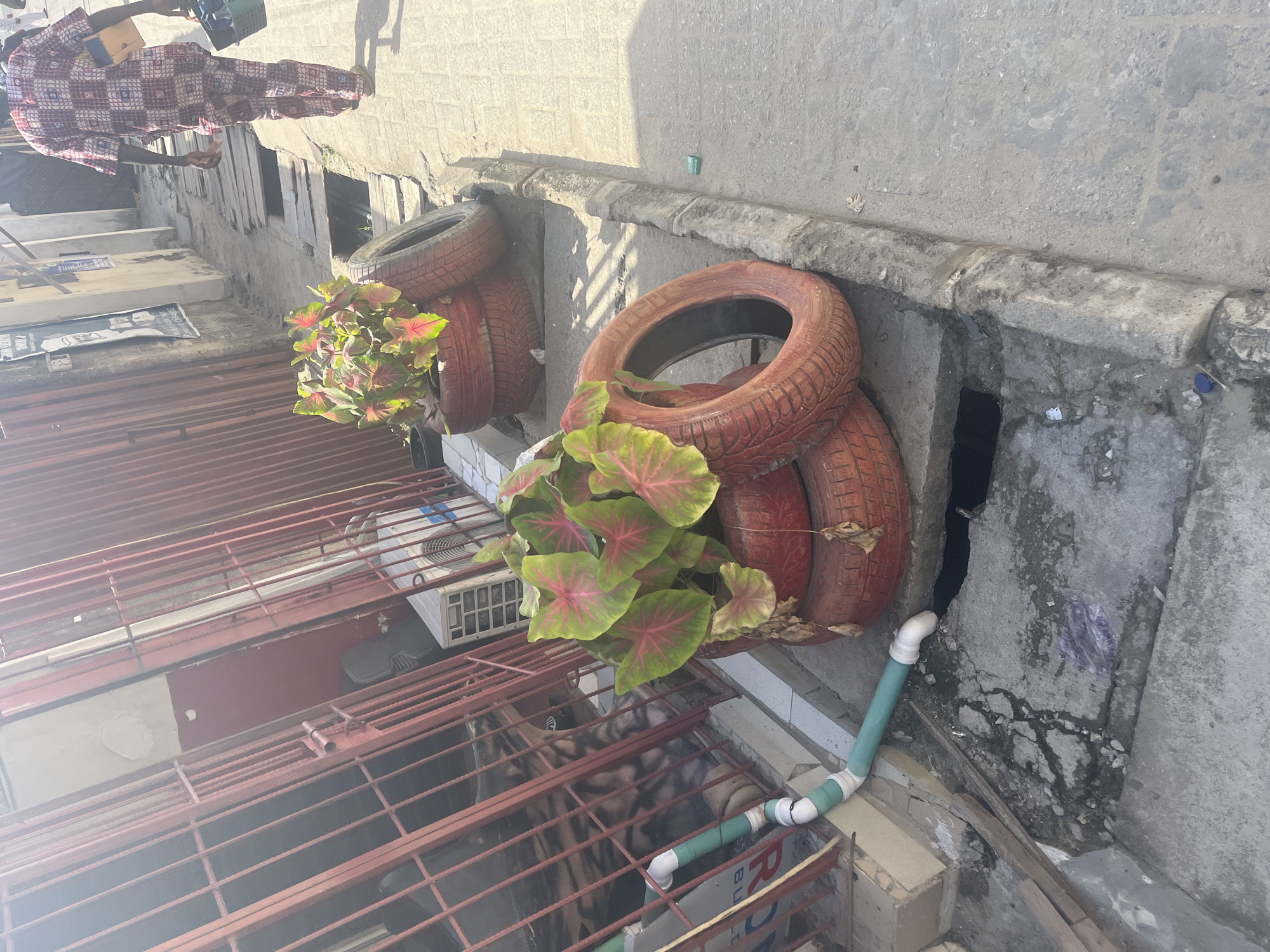 The image shows an exterior scene in Ado, Nigeria, likely a commercial or residential area. Viewed from above, a red iron grate overlooks a courtyard or walkway below. Two sets of old tires painted red are stacked to form planters, each containing a leafy plant. The tires are situated against a weathered concrete wall, possibly bordering a street or alley. A blue and white plastic pipe runs along the base of the wall. Part of a person wearing a long-sleeved, patterned shirt is visible in the upper left corner of the frame, near the building entrance.
