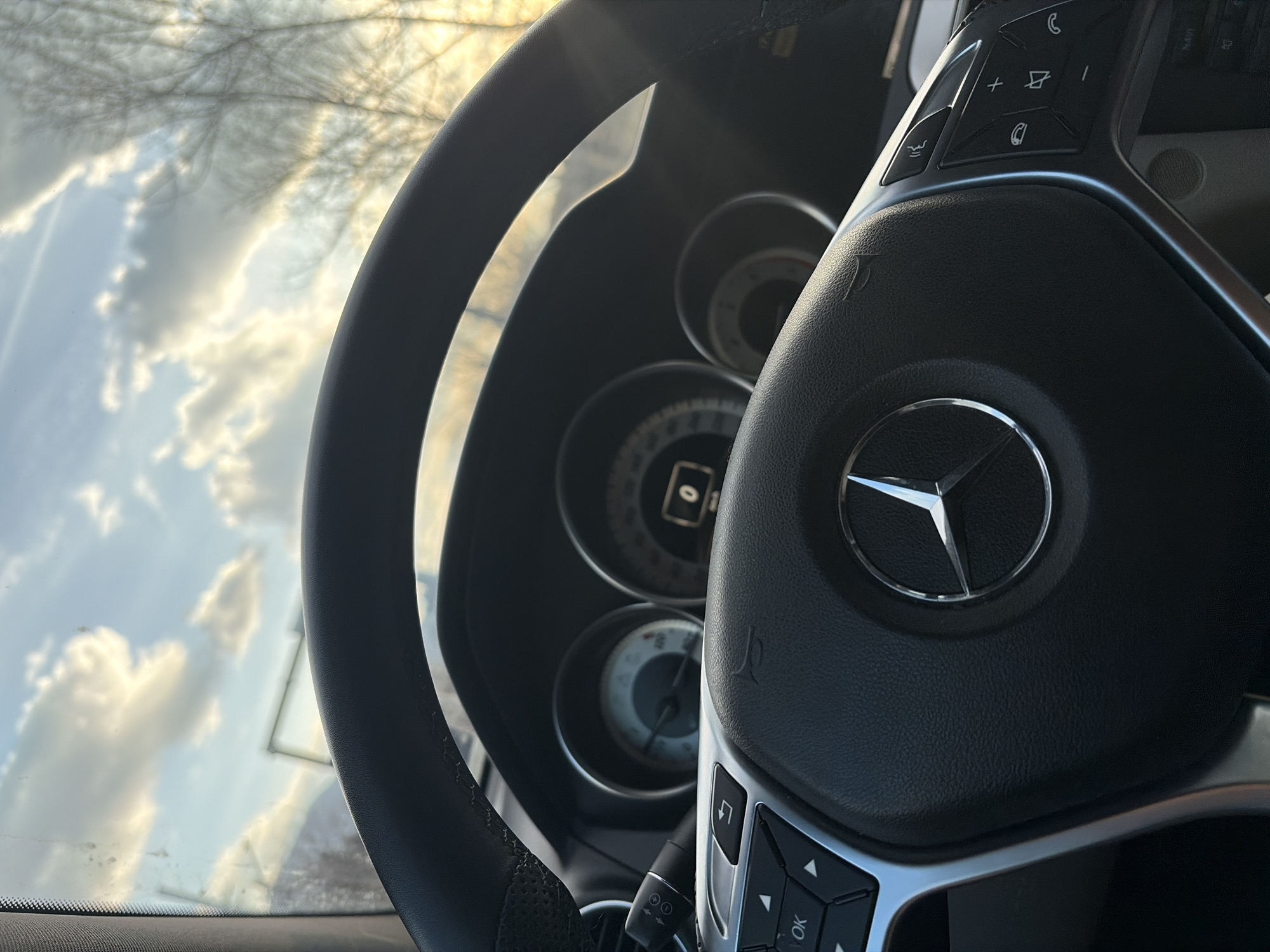 The image presents a forward-facing view from inside a Mercedes-Benz vehicle, located in Sofia, Bulgaria, during what appears to be late afternoon or early evening. The dominant subject is the vehicle's black, leather-wrapped steering wheel, featuring a prominent chrome Mercedes-Benz three-pointed star emblem in its center. The steering wheel includes silver-toned multi-function controls on both the left and right spokes, with buttons for navigation (directional arrows, "OK") on the left and telephone/audio controls (call, hang up, volume) on the right.
Behind the steering wheel, a portion of the instrument cluster is visible, showing two circular analog gauges and a central digital display within the right gauge indicating "0". A control stalk is partially visible on the left side, extending from the steering column. Through the vehicle's windshield, the sky is visible, characterized by scattered white and grey clouds against a light blue backdrop. The sun, positioned low, casts a golden hue, particularly towards the right side of the frame. Bare tree branches are silhouetted against this bright sky on the right side, suggesting a deciduous tree without leaves. Minor dirt or streaks are visible on the upper left portion of the windshield. No individuals are directly visible in the frame.